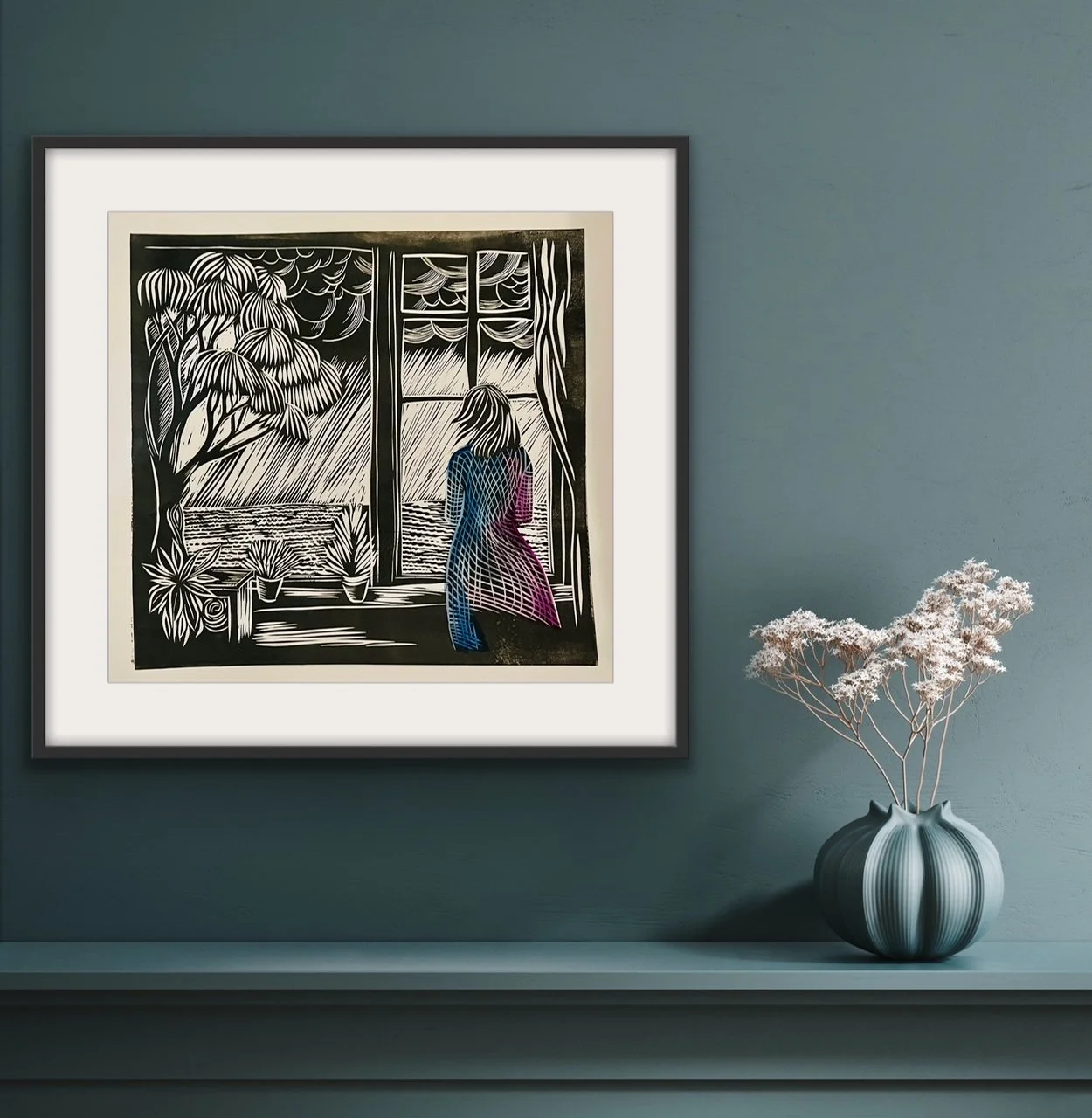 Artwork of a woman looking out a window with plants, trees, and a cloudy sky outside, framed with a black border on a wall with a console below and a white flower arrangement in a striped vase.