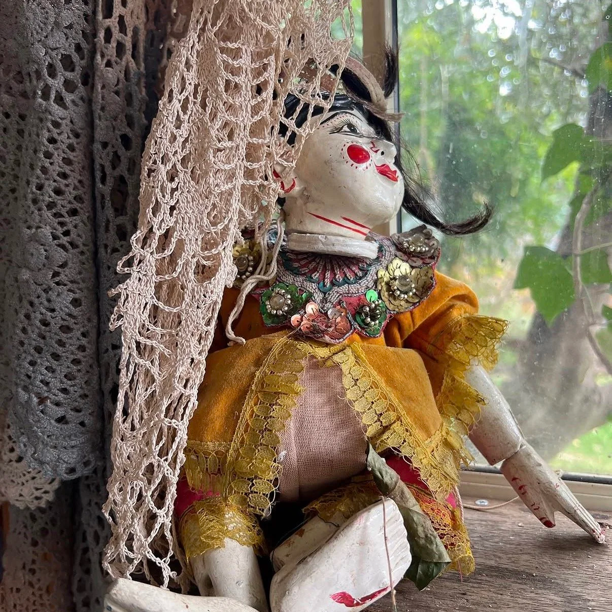 A vintage handcrafted doll with painted face, black hair, and traditional clothing, sitting on a wooden surface near a window with greenery outside.