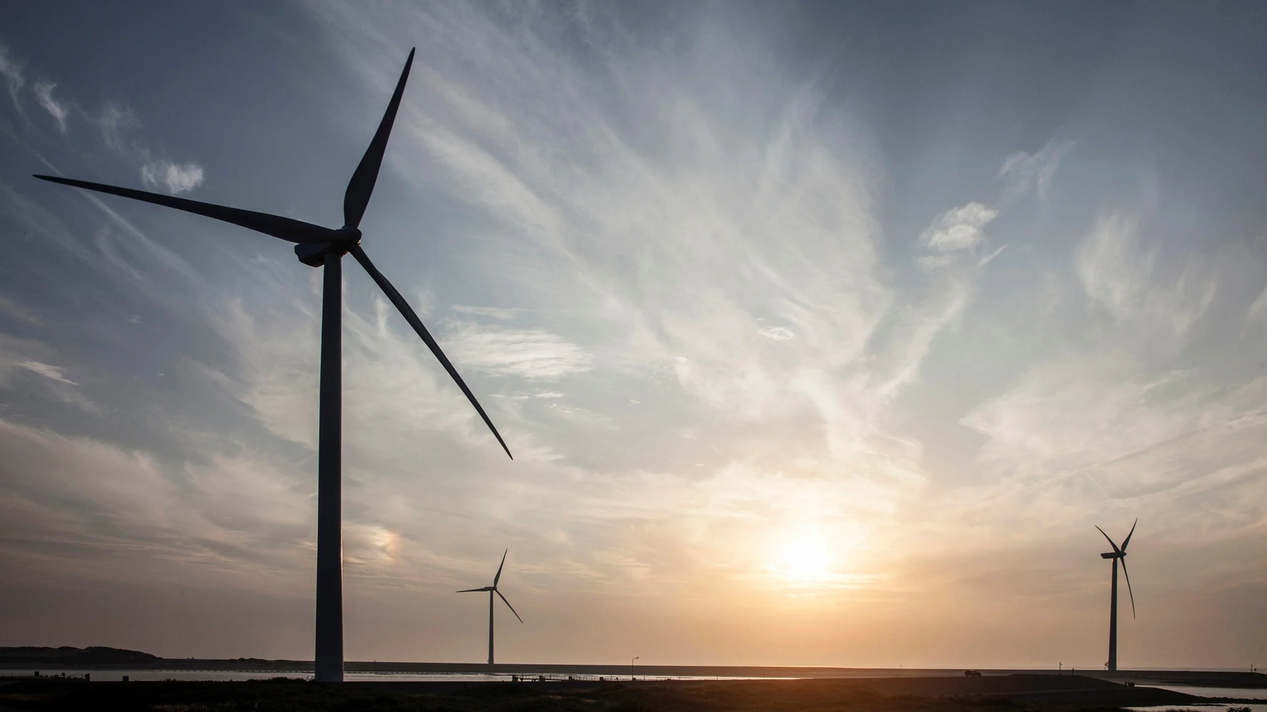 Several wind turbines on a flat landscape during sunset with a partly cloudy sky.