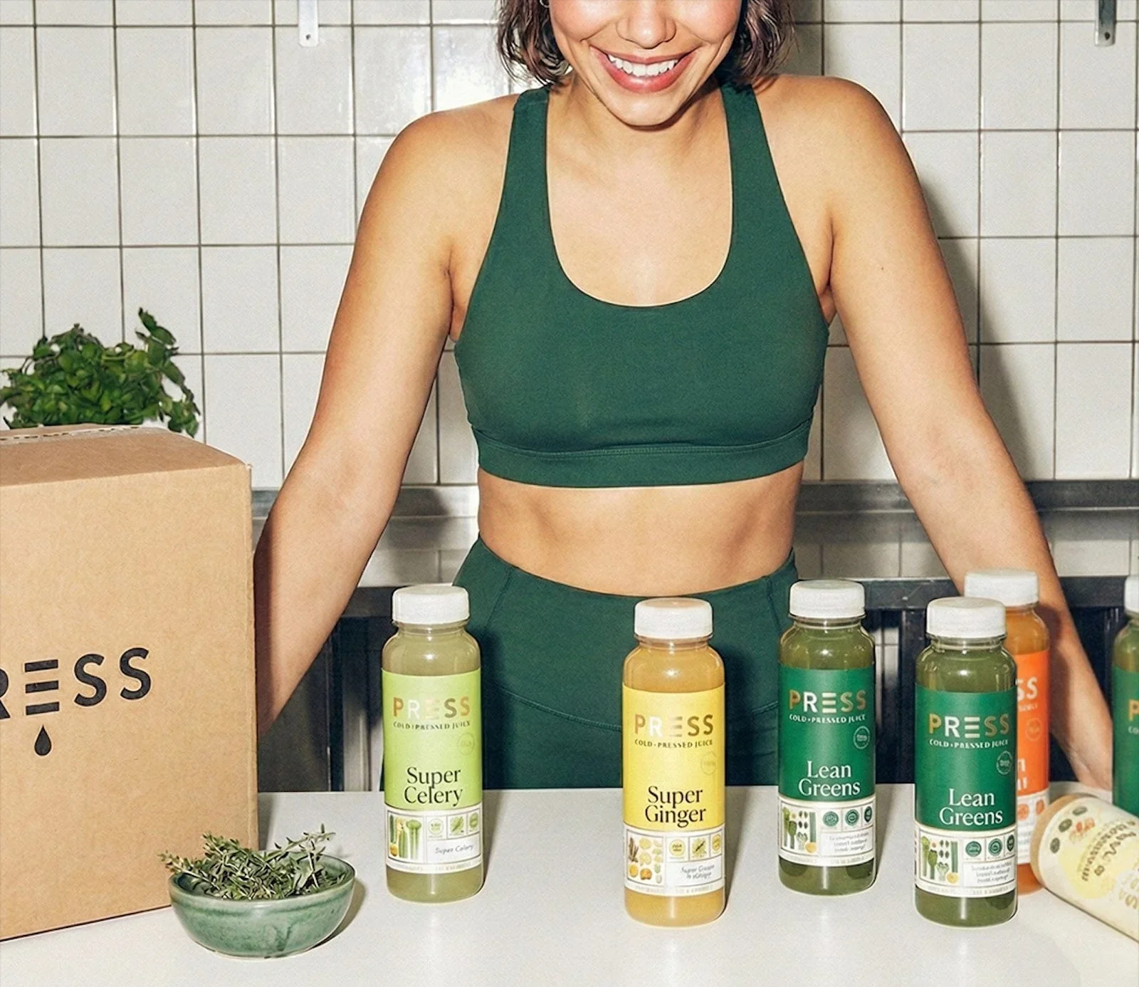 Woman smiling at a juice tasting table with bottles labeled Super Celery, Super Ginger, and Lean Greens, and a small potted herb in front.