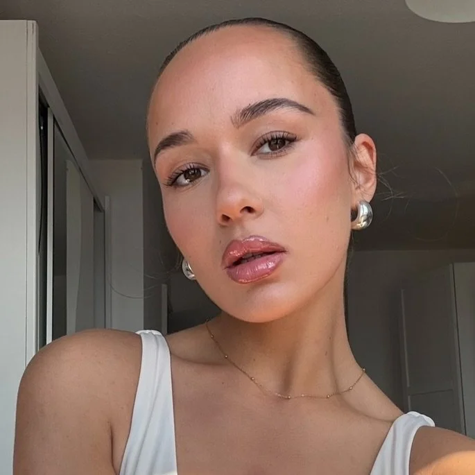Close-up of a woman with pulled-back hair, wearing pearl earrings and a white top, in a well-lit room.