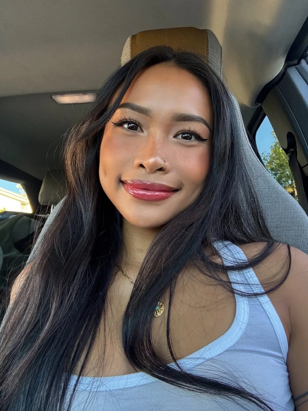 A young woman with long dark hair, wearing makeup, a silver nose ring, and a white tank top, sitting in a car, smiling at the camera.