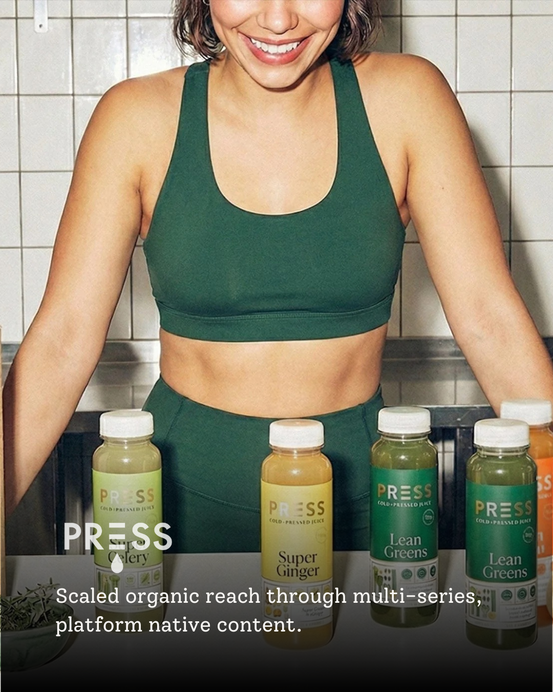 A woman in a green sports bra standing behind a table with four bottles of juice labeled 'PRESS'. The bottles are labeled 'Super Ginger', 'Lean Greens', and 'Celery'. There is a bowl of herbs in the lower left corner.