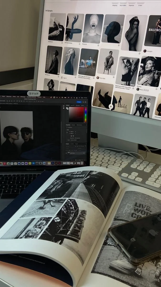 A workspace with a desktop monitor displaying a Pinterest page with black and white fashion photos, a laptop showing a photo editing program with two portraits of women, an open magazine with black and white images and text, a smartphone, and a white keyboard.