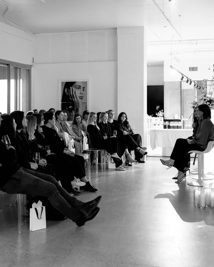 A woman is speaking on a stage in front of an audience of women in a modern, minimalist space.