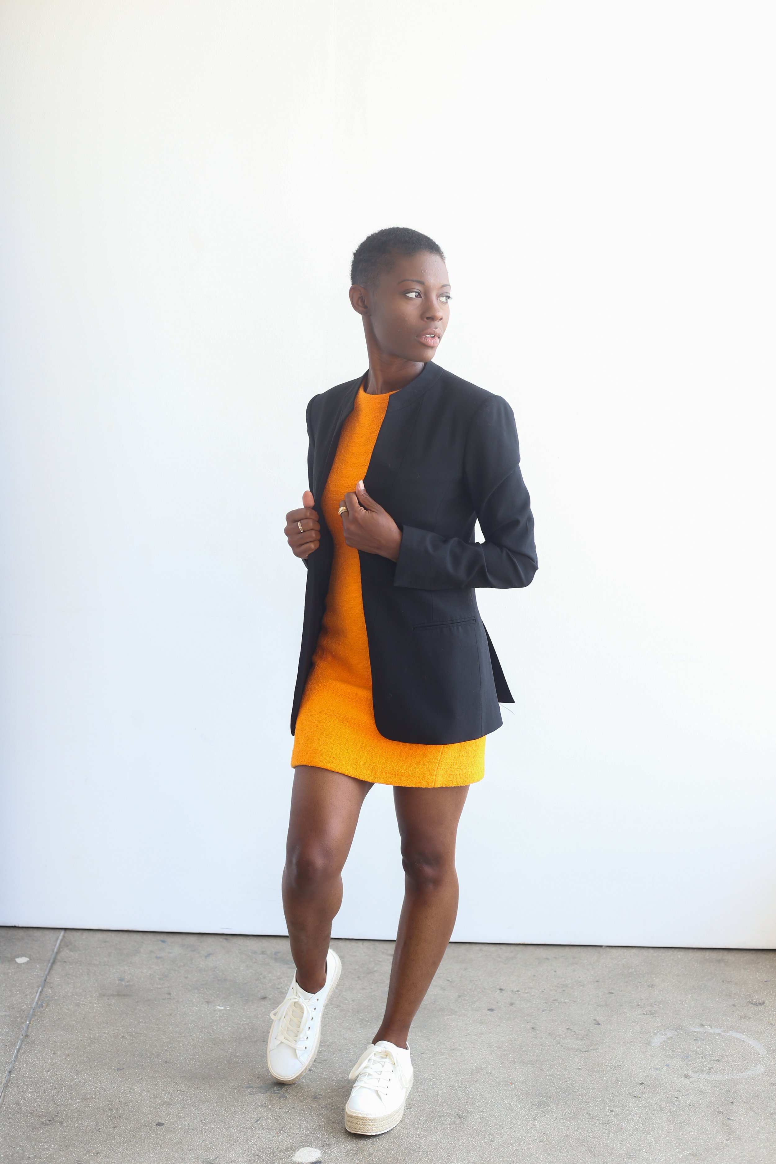 A woman standing against a plain white wall, wearing a yellow dress, a black blazer, and white sneakers, looking to her right.