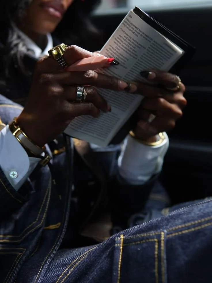 Person reading a guidebook or magazine inside a car, wearing jewelry and denim clothing.