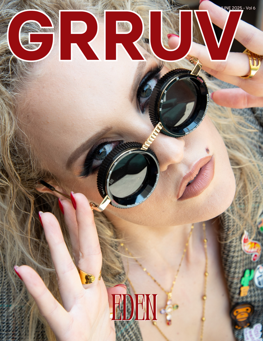 Close-up of a woman with curly Blonde hair wearing stylish sunglasses with dark lenses and gold accents, holding the glasses near her face. She has light makeup with nude lipstick, small piercings, and colorful pins on her jacket. She also wears gold
