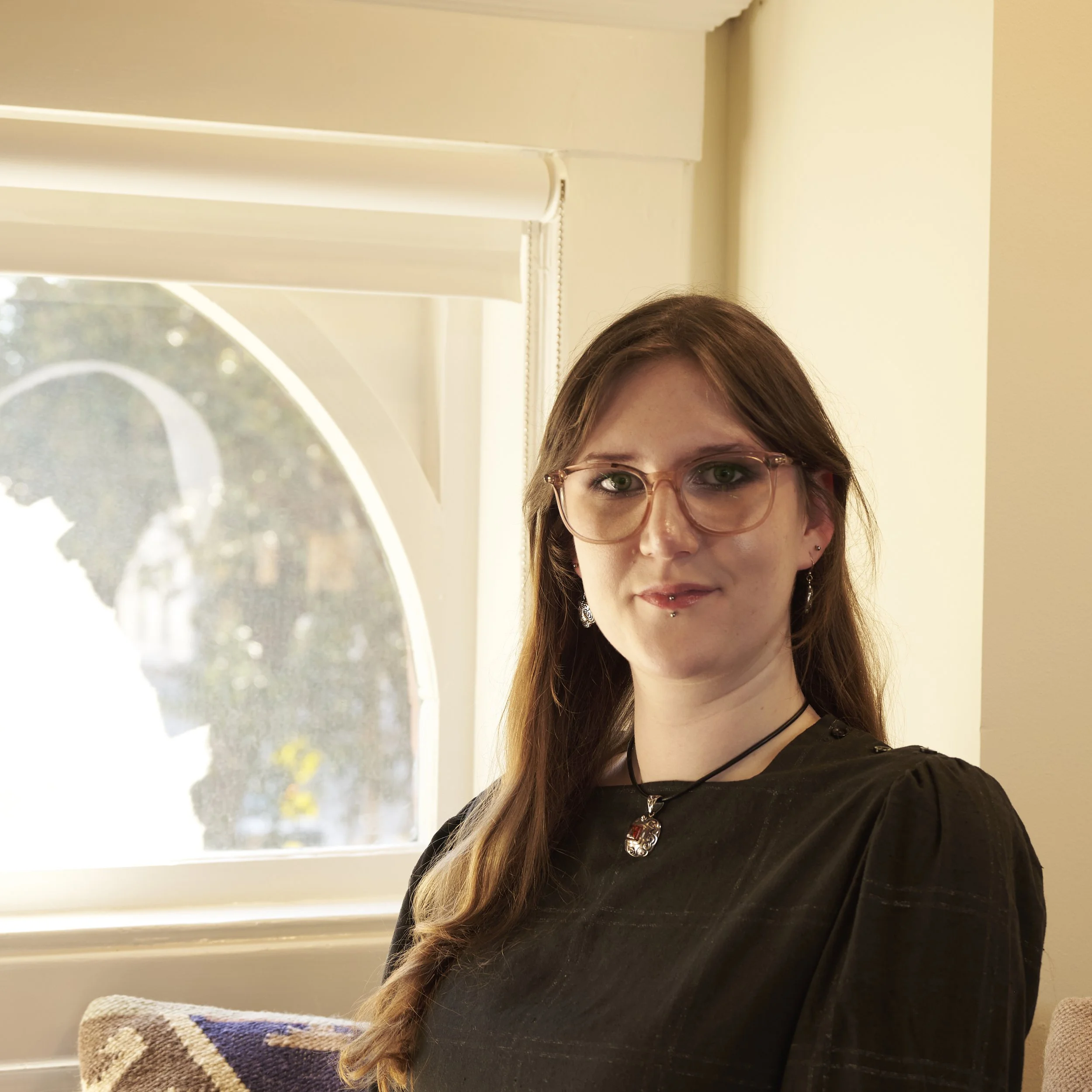 A woman with long brown hair, glasses, and piercings, wearing a black top with jewelry, sitting near a window with a blurred outdoor background.