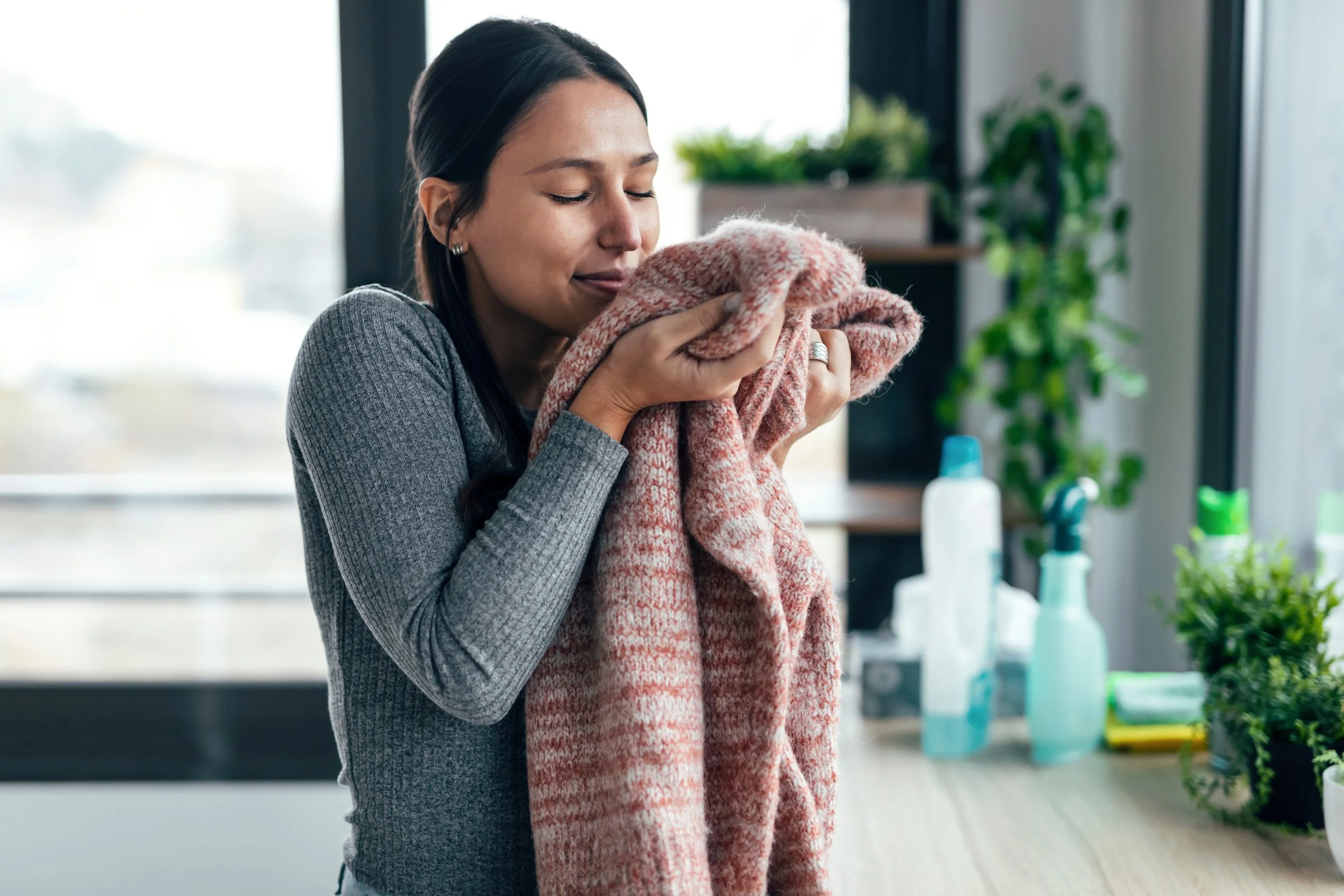 A woman smelling a pink and beige patterned scarf indoors, near a window with plants and cleaning supplies on a counter.