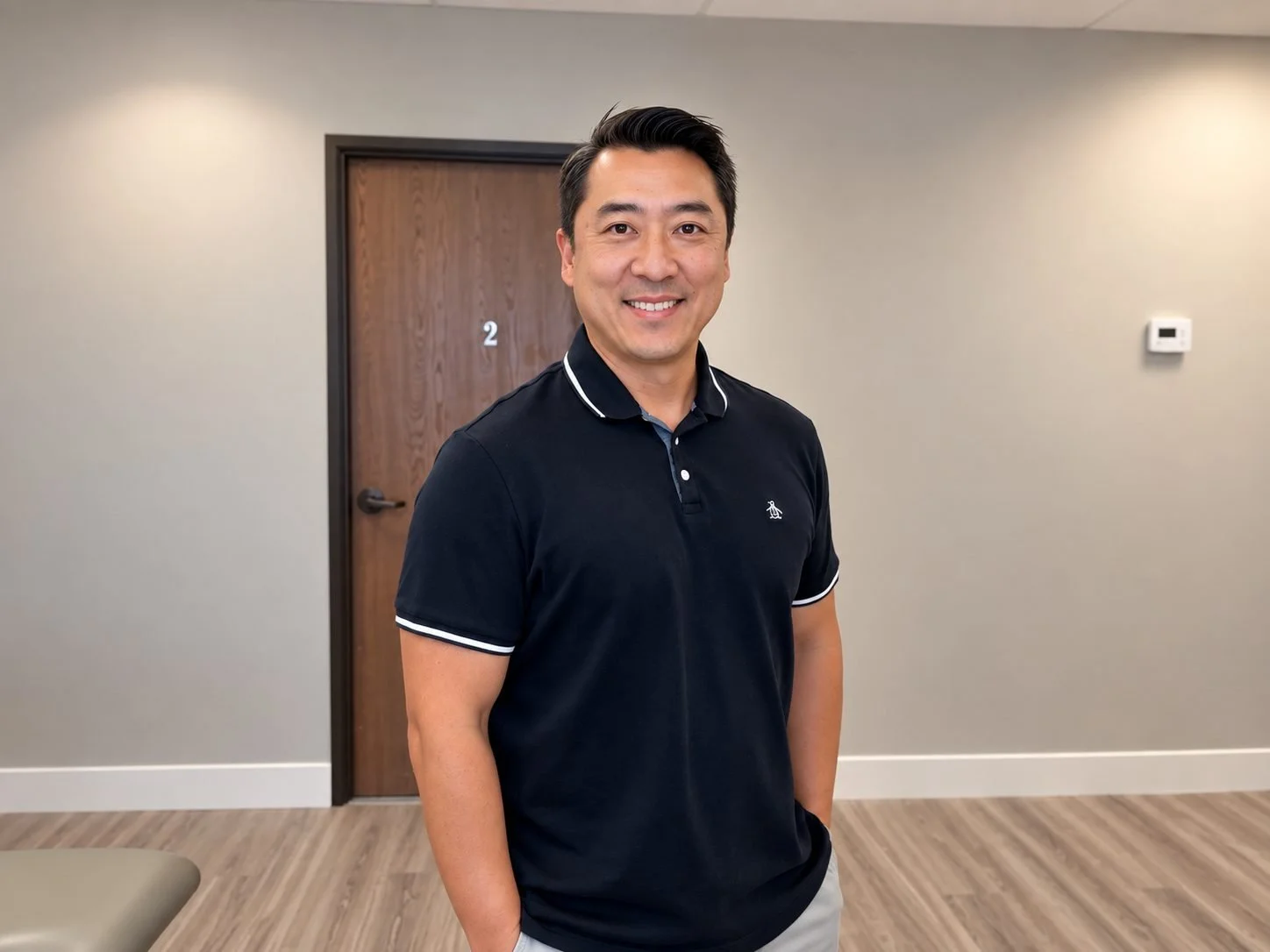 A man smiling and looking at the camera, standing indoors near a wooden door with the number 2 on it. He is wearing a black polo shirt with white accents.