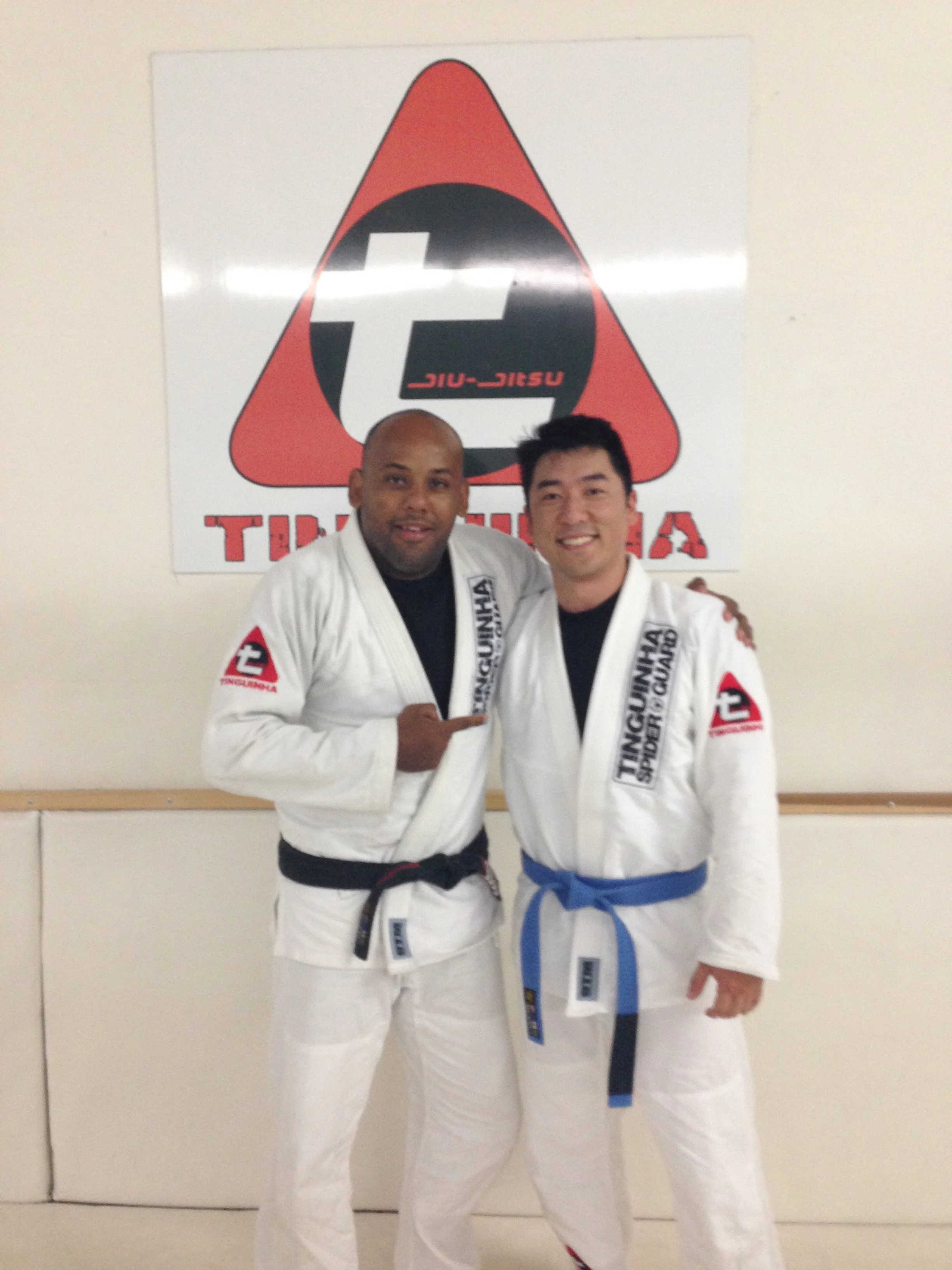 Two men in white martial arts uniforms, one with a black belt and one with a blue belt, standing in front of a logo for Tinguinha Brazilian Jiu-Jitsu, smiling and posing for a photo.