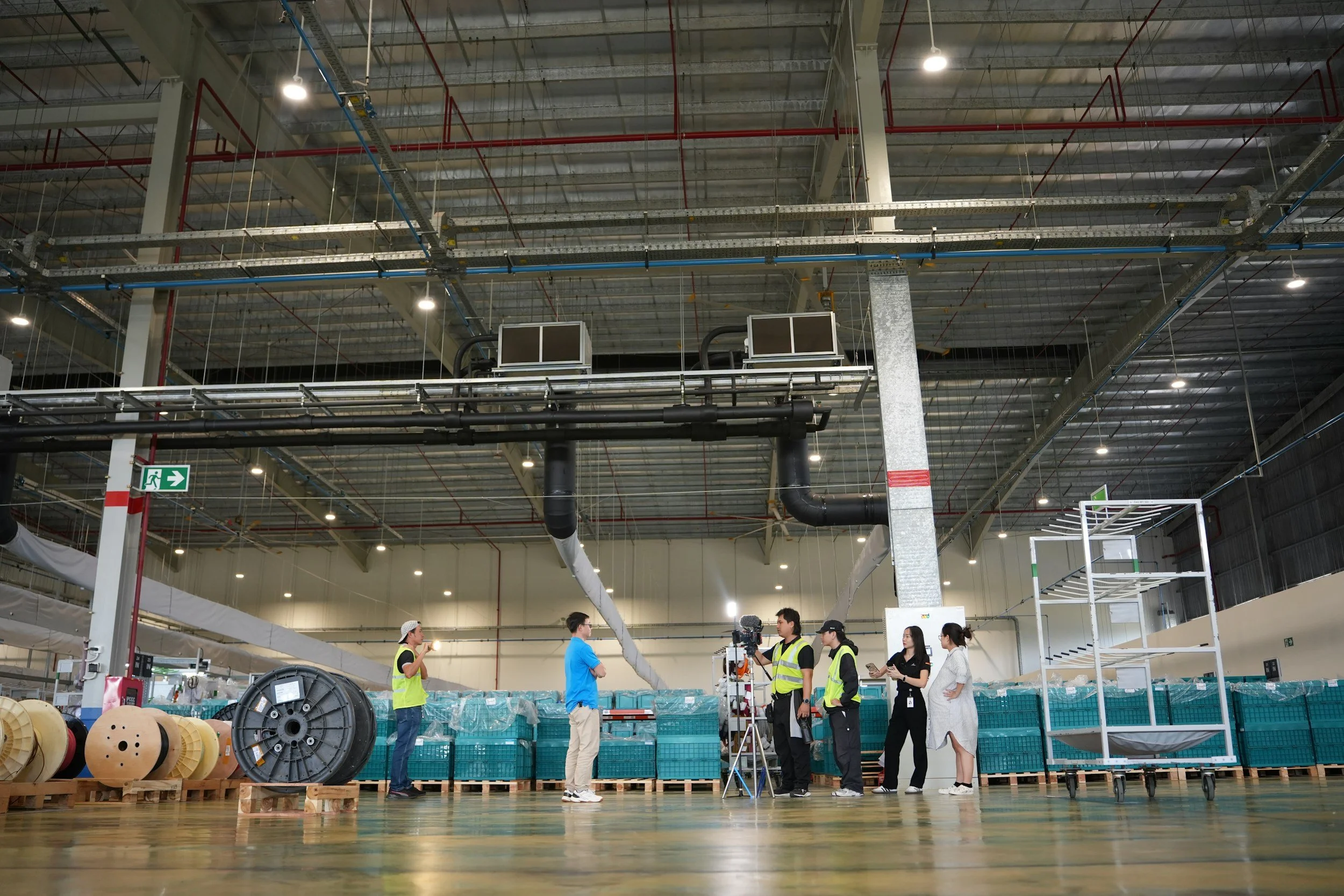 Group of people in safety vests and casual clothing in a large industrial warehouse, with equipment, spools, and scaffolding around, likely filming or inspecting.