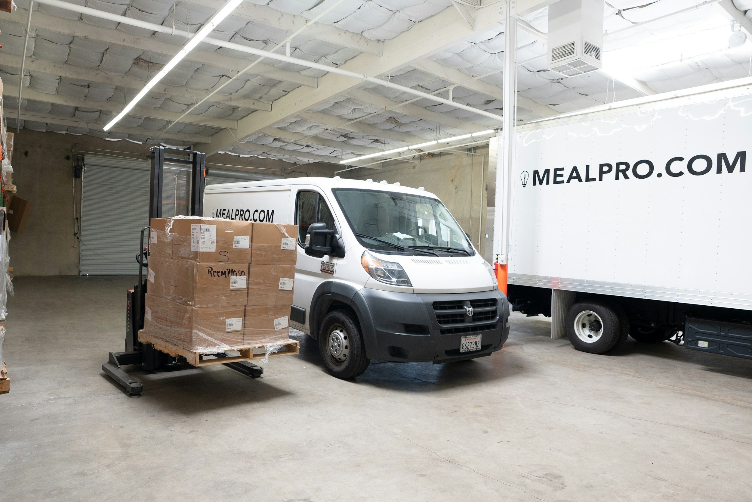 A white delivery van and a large white cargo truck with 'MEALPRO.COM' written on their sides are parked inside a warehouse, with a pallet of boxed goods on a hand truck nearby.