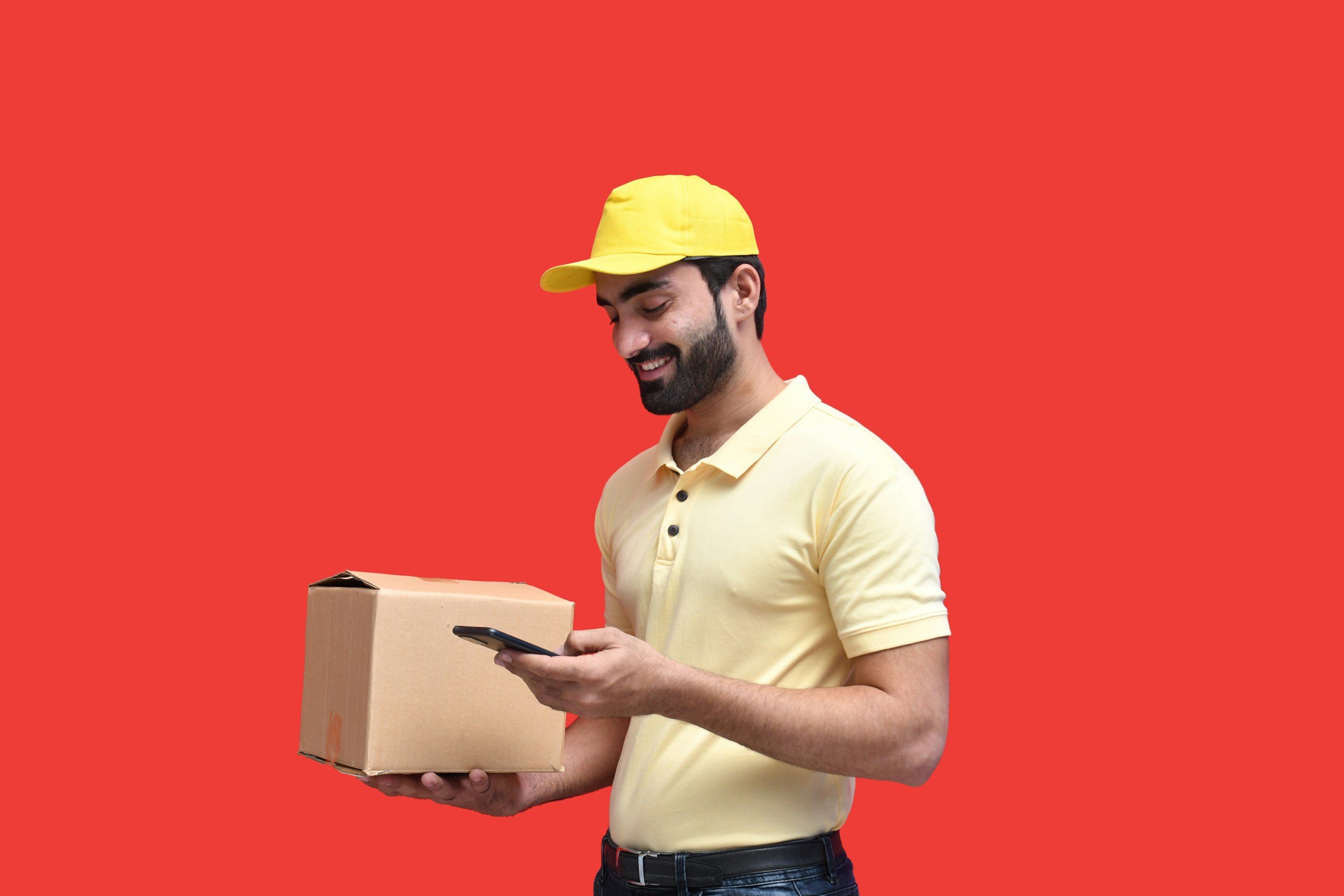Delivery person in yellow cap and shirt holding a cardboard box while using a smartphone against a red background.