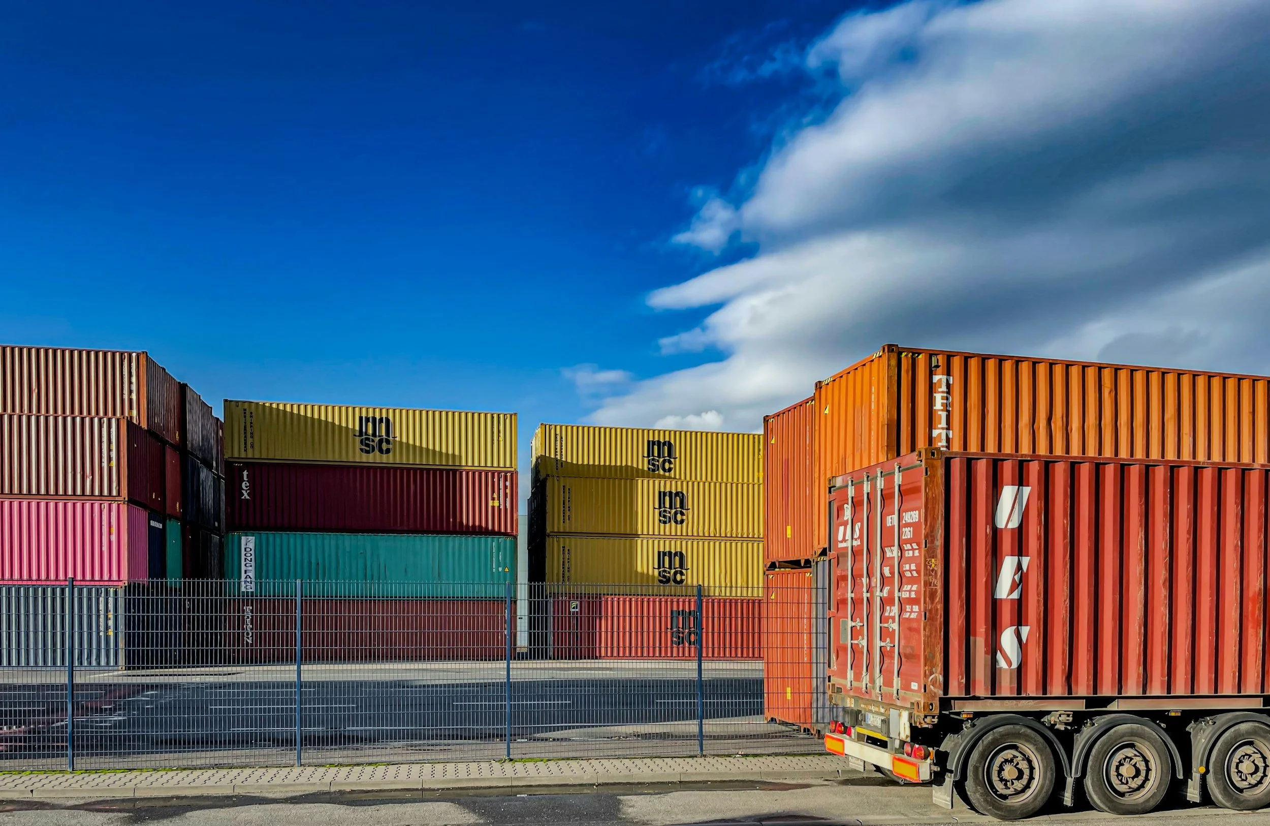Colorful stacked shipping containers behind a metal fence, with a blue sky and clouds overhead.