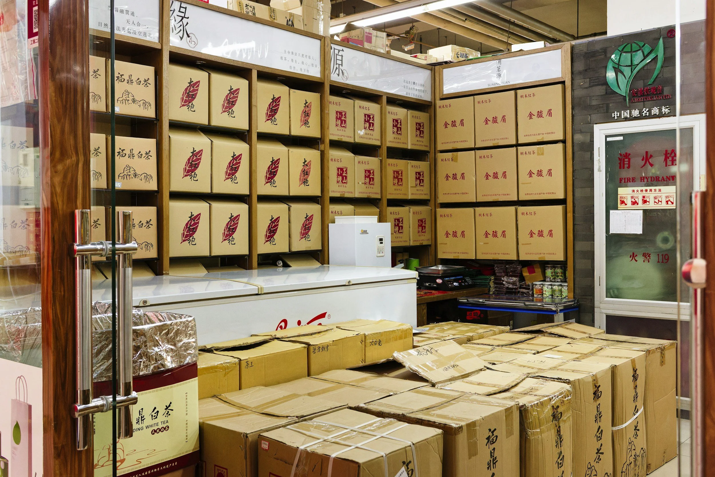 Store with shelves filled with cardboard boxes labeled in Chinese, some stacked on the floor, and a chest freezer in the front. A fire hydrant cabinet is on the right side.