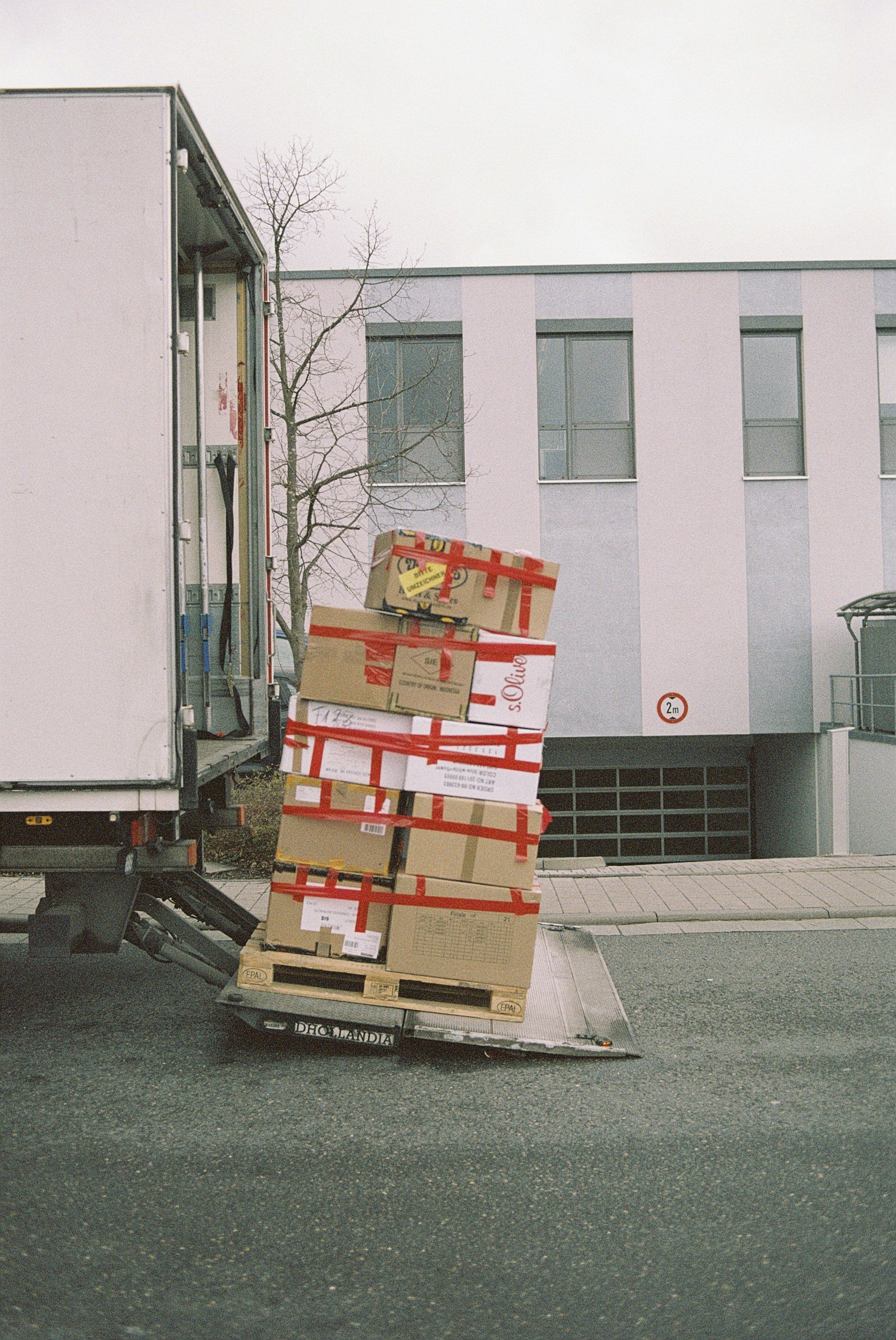Delivery truck with stacked cardboard boxes on a luggage cart outside a modern building.