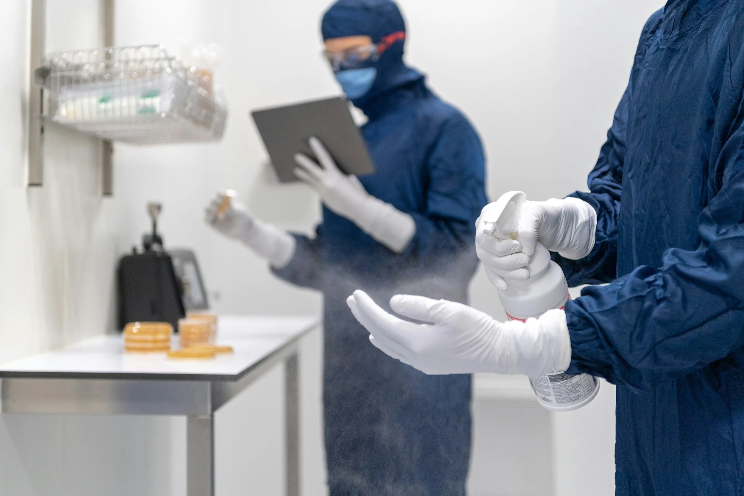 Two people in protective gear working in a laboratory, one using a spray bottle and the other handling a tool, with petri dishes on the table.