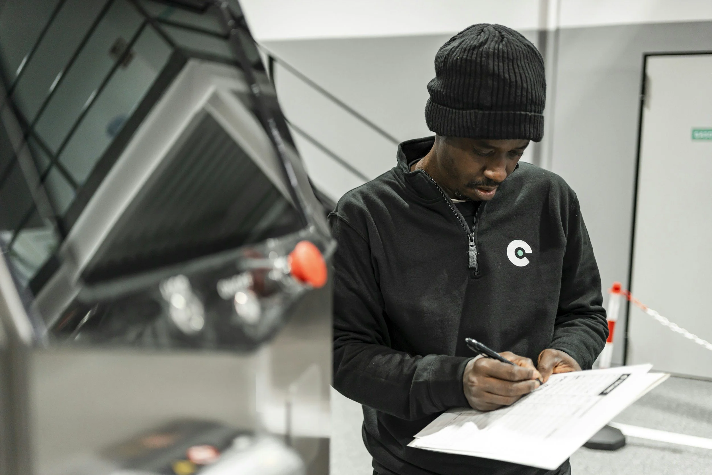 A man wearing a black beanie and black jacket with a logo, writing on a clipboard in an industrial or hospital setting.