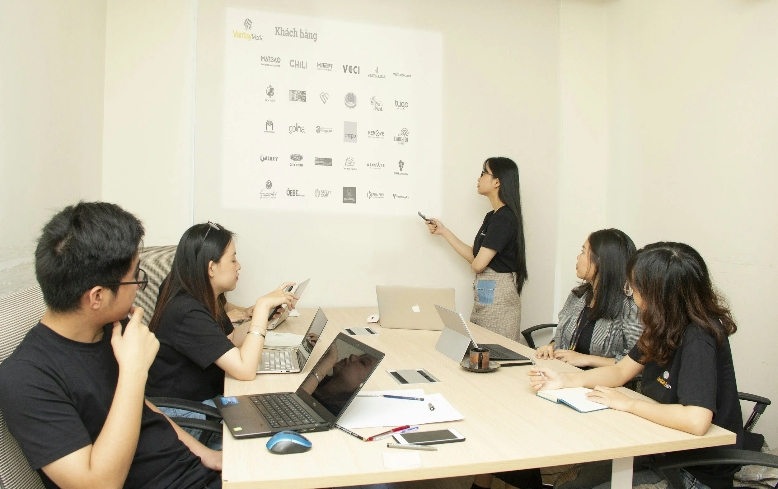 A woman presents to four seated colleagues during a business meeting in a conference room with laptops and notebooks on the table.