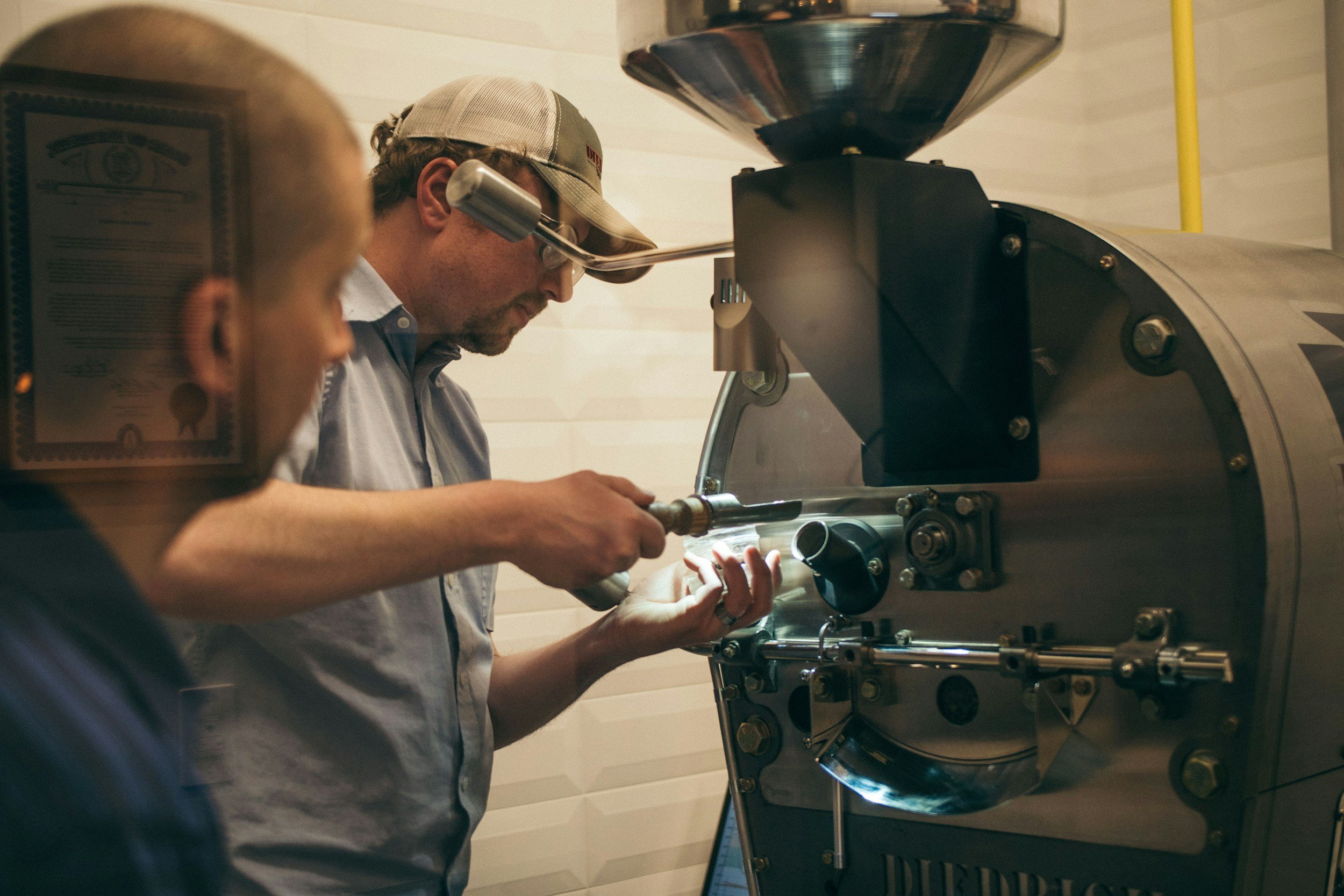 Two men are working with a large coffee roaster machine, one is inspecting or adjusting something on the machine while the other observes.
