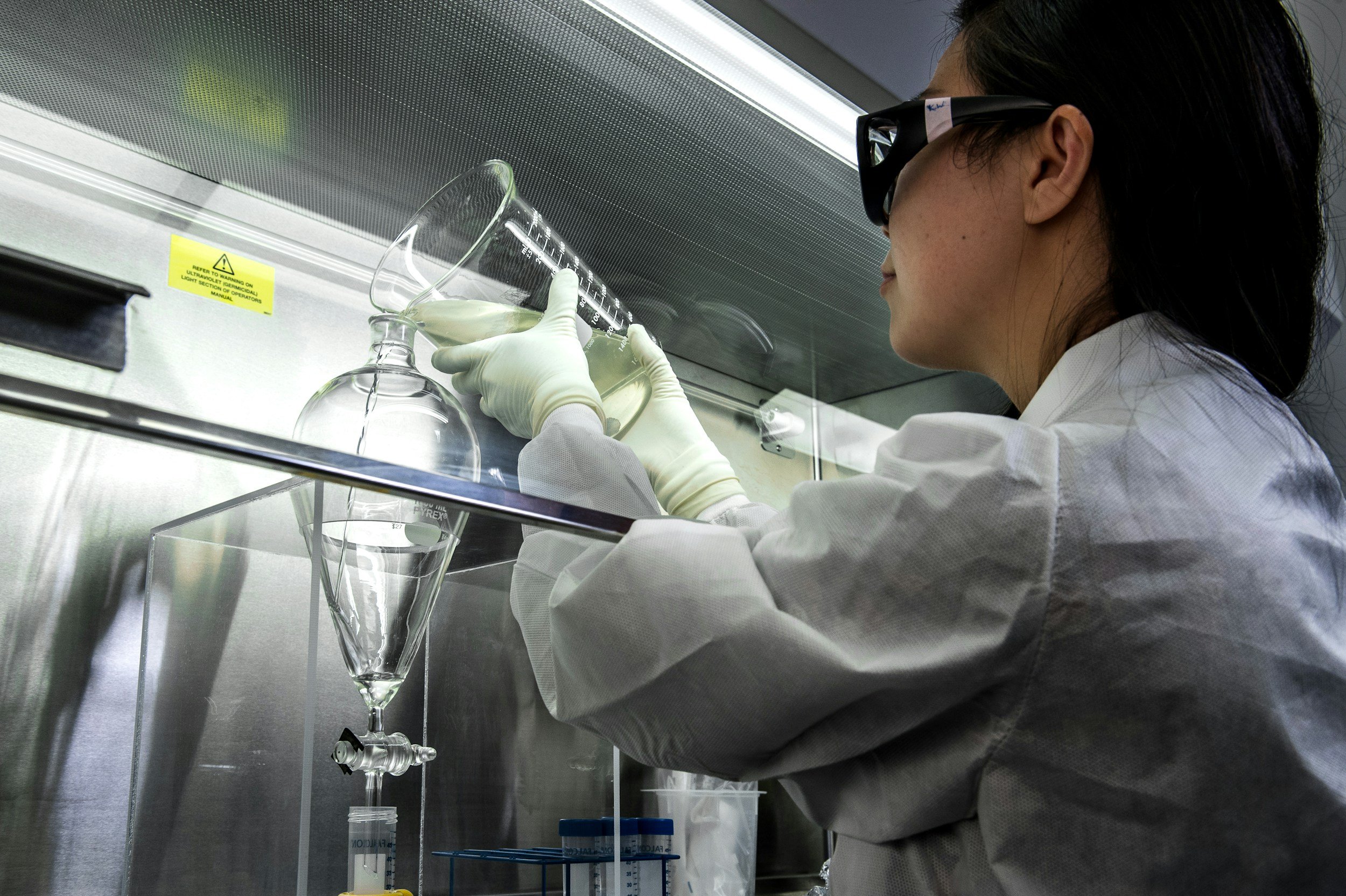 A scientist wearing safety glasses, gloves, and a lab coat is working in a laboratory. She is using a funnel to transfer a clear liquid into a glass bottle inside a fume hood.