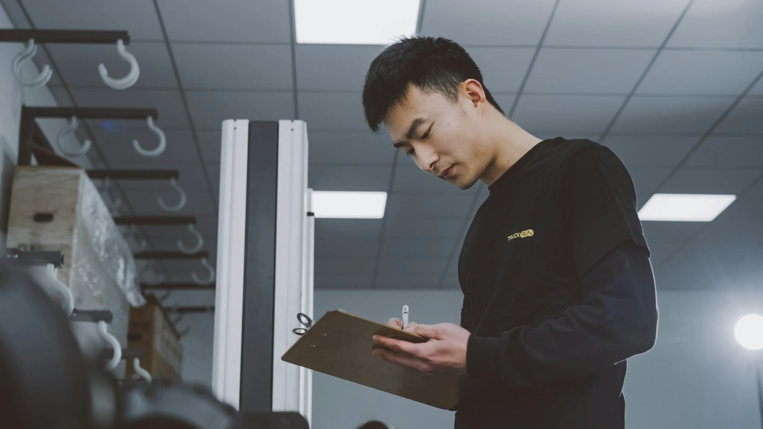 A young man in black clothing, writing on a clipboard in a gym or fitness studio.