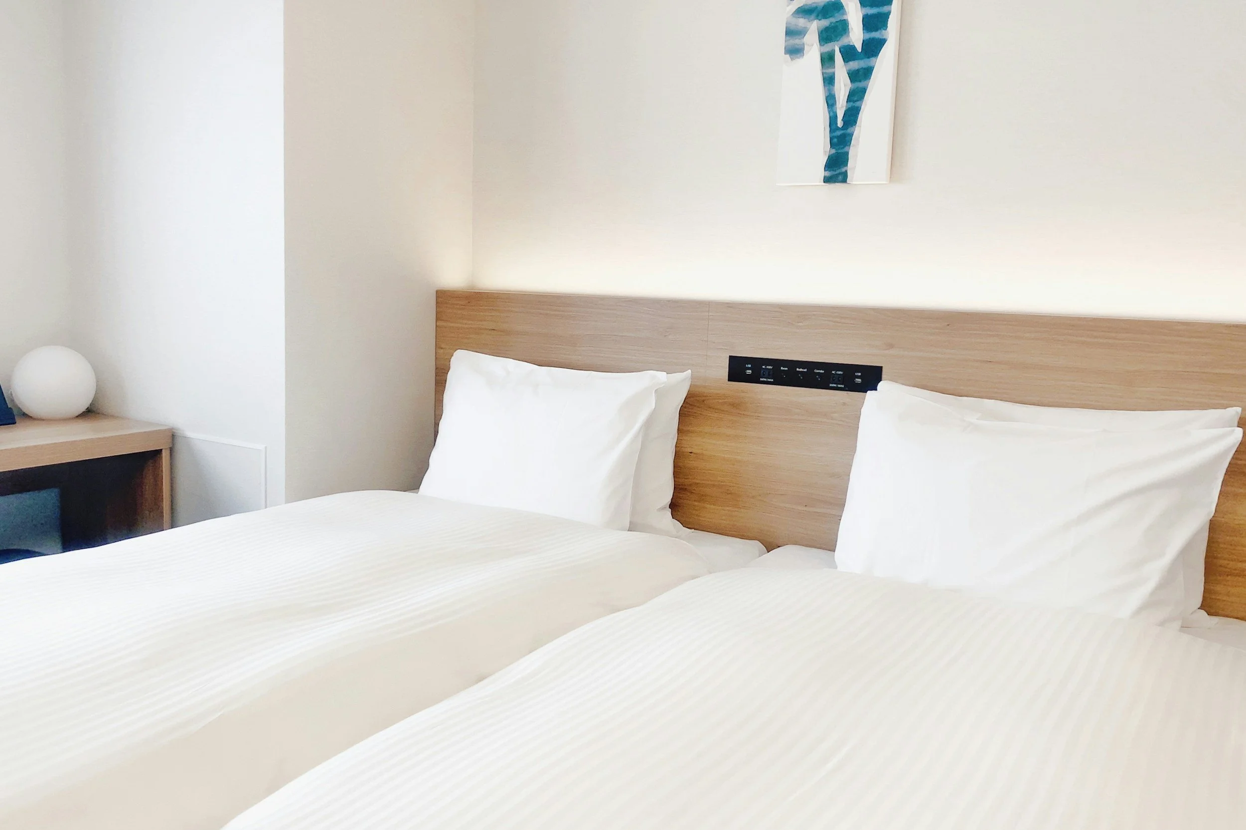 Two neatly made hotel beds with white linens and pillows, wooden headboards, in a modern minimalistic room with a nightstand, a decorative lamp, and an abstract painting on the wall.