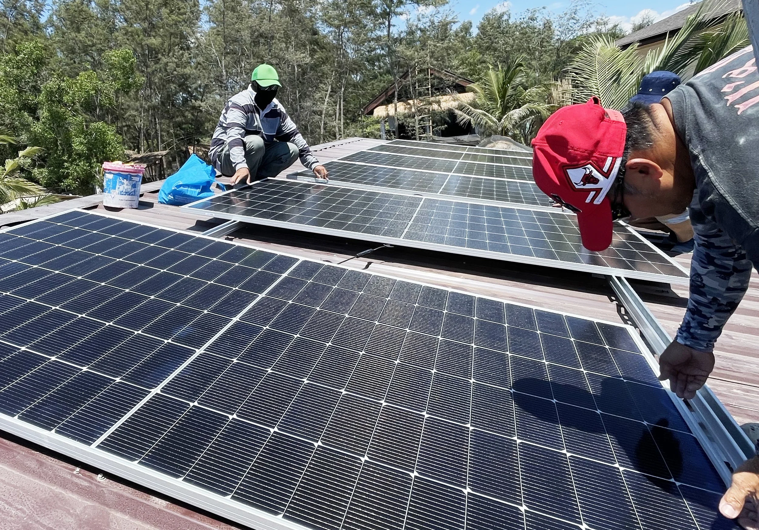 Two workers installing solar panels on a rooftop in a sunny outdoor setting with trees and houses in the background.