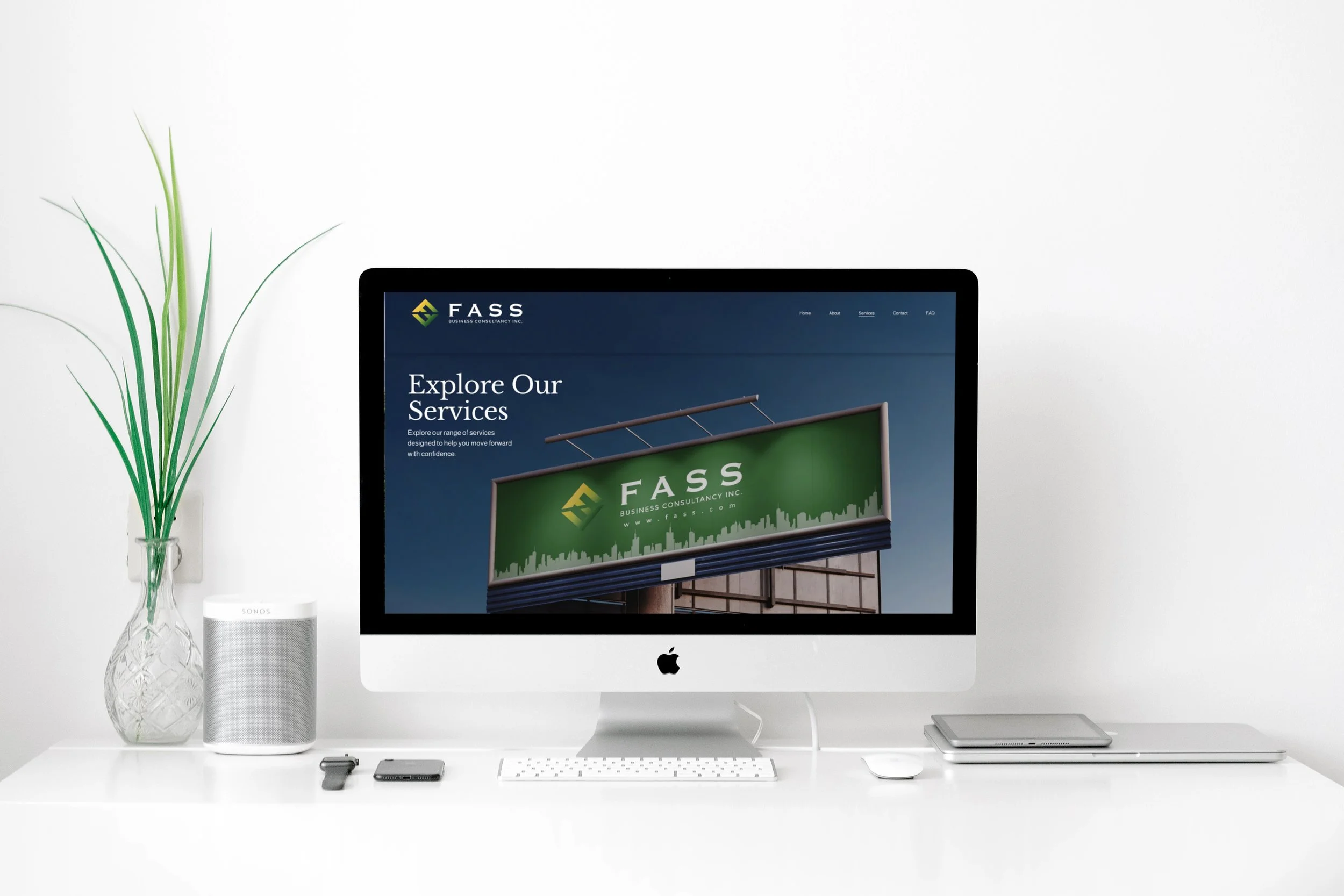A white desk with an iMac displaying a website for FASS Business Consultancy Inc., with the text "Explore Our Services" on the screen. The desk has a potted green plant, a white speaker, a smartphone, a smartwatch, and a closed laptop.