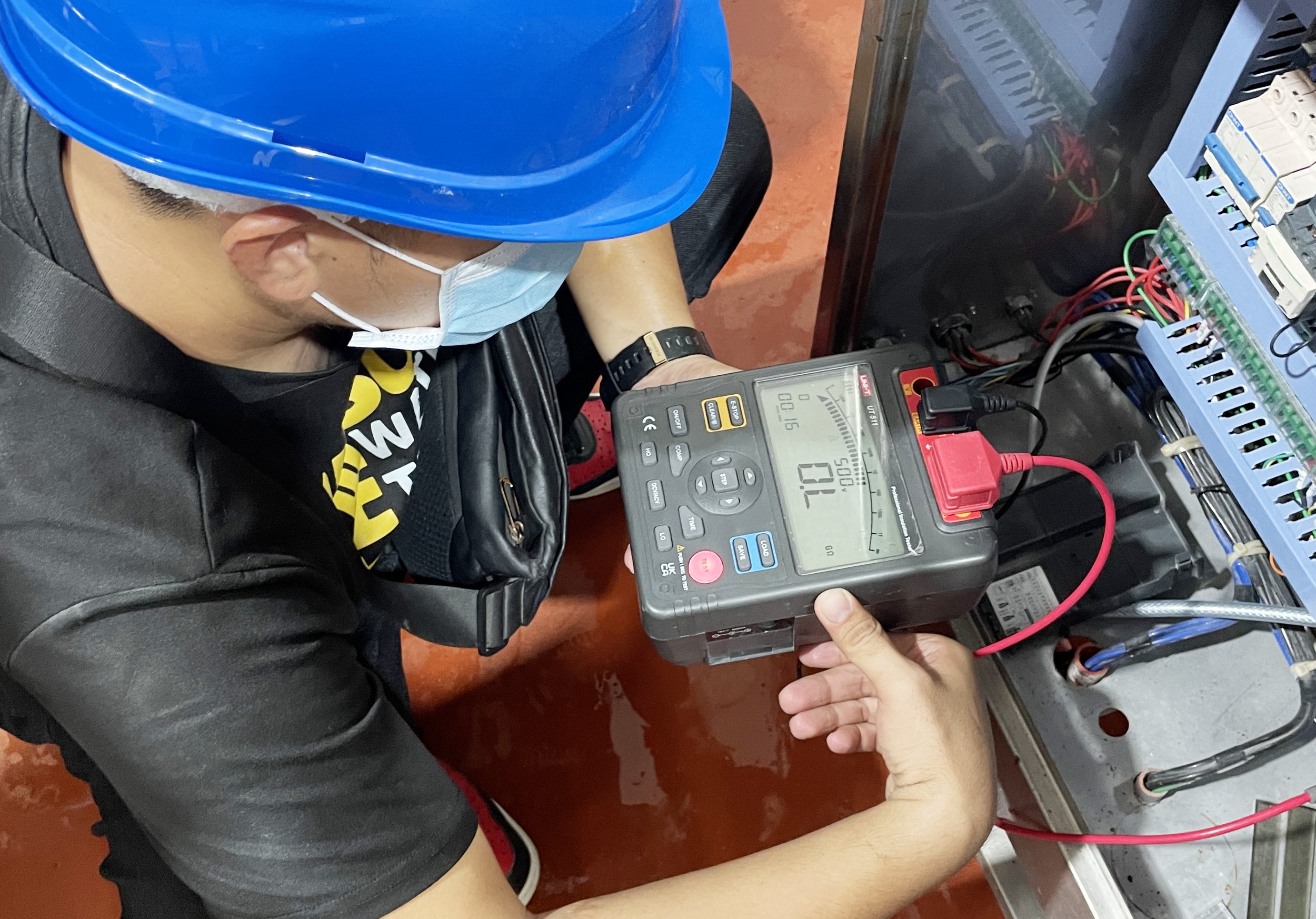 Worker wearing a blue safety helmet and face mask testing electrical circuit with a digital multimeter in a control panel.