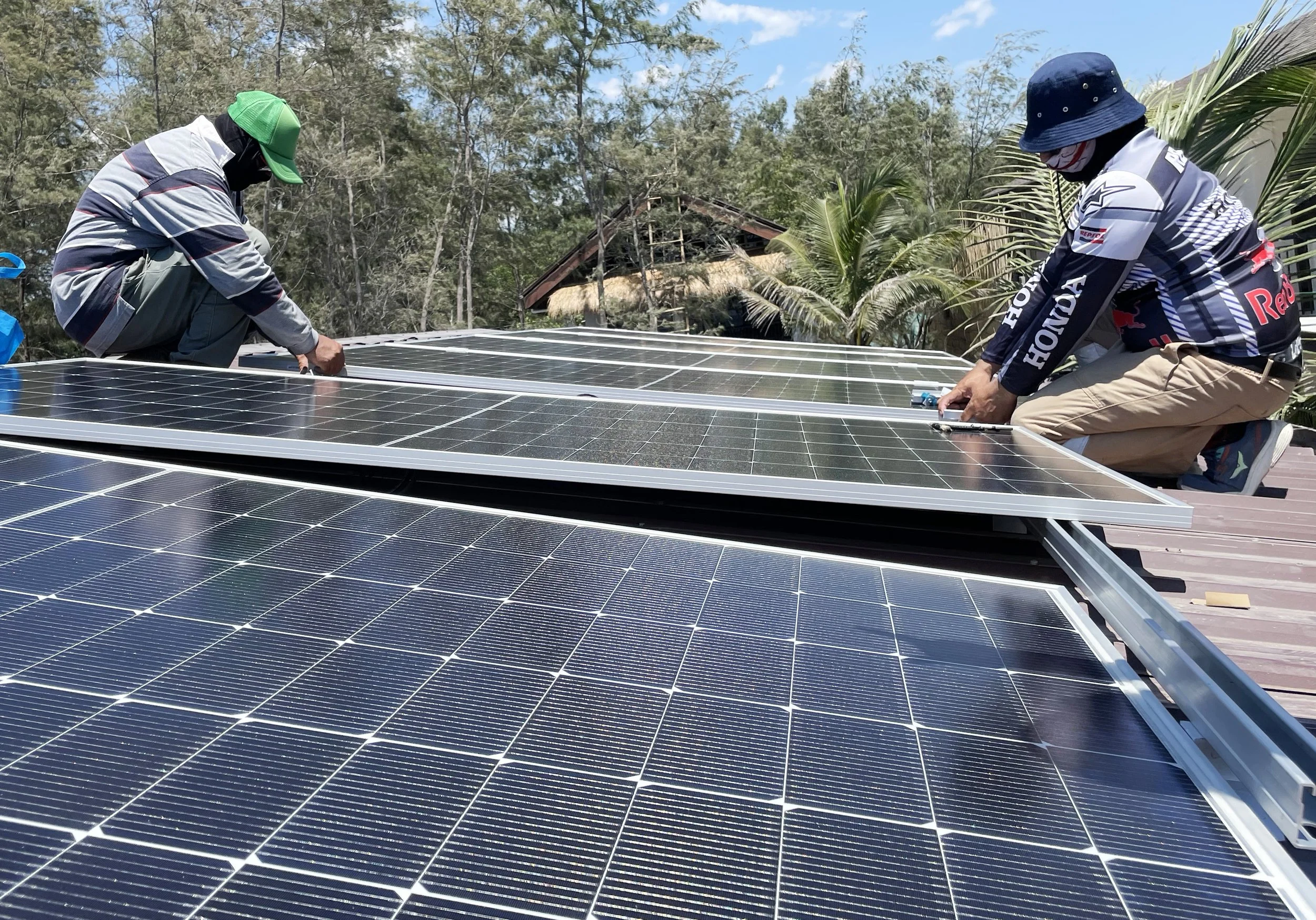 Two people working on a rooftop solar panel installation, with trees and a house in the background during daytime.
