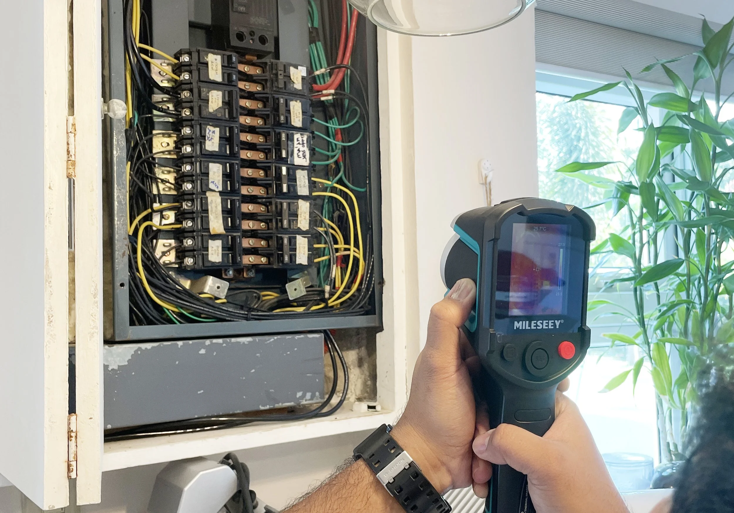 Person using a thermal imaging camera to inspect an electrical breaker panel with multiple switches and wires.