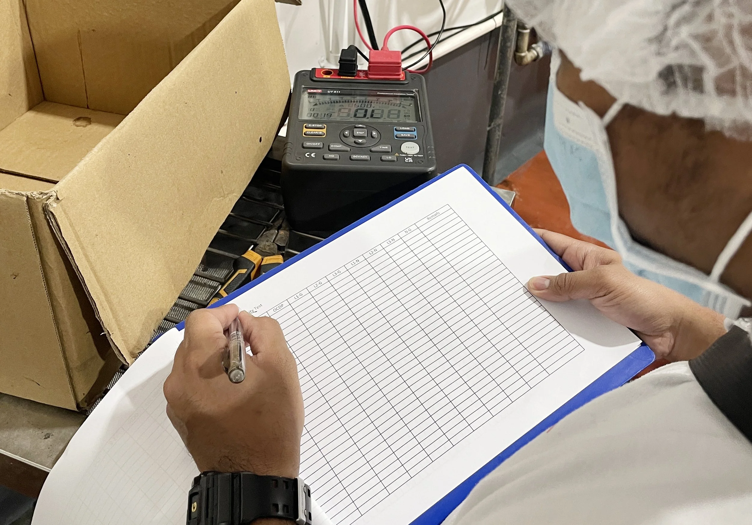 Person wearing a face mask, holding a checklist or record sheet, and writing on it with a pen. There is a digital device, possibly a multimeter or measuring instrument, placed on a cart, along with a cardboard box and some tools.