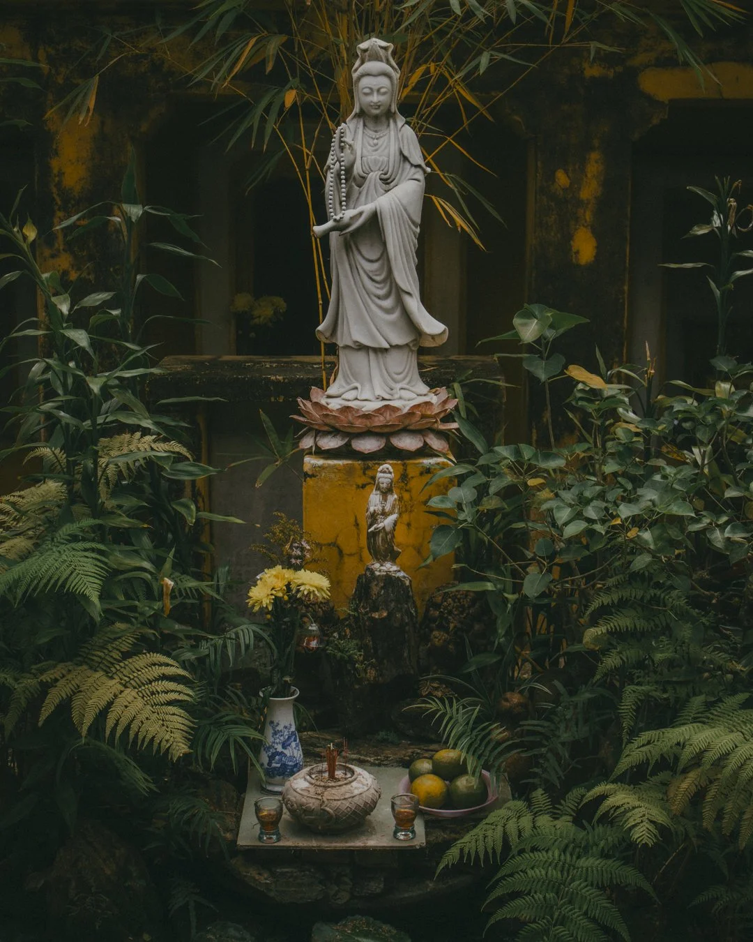 A statue of a serene standing deity surrounded by lush green plants and flowers, with offerings like flowers, fruits, and candles in front of it.