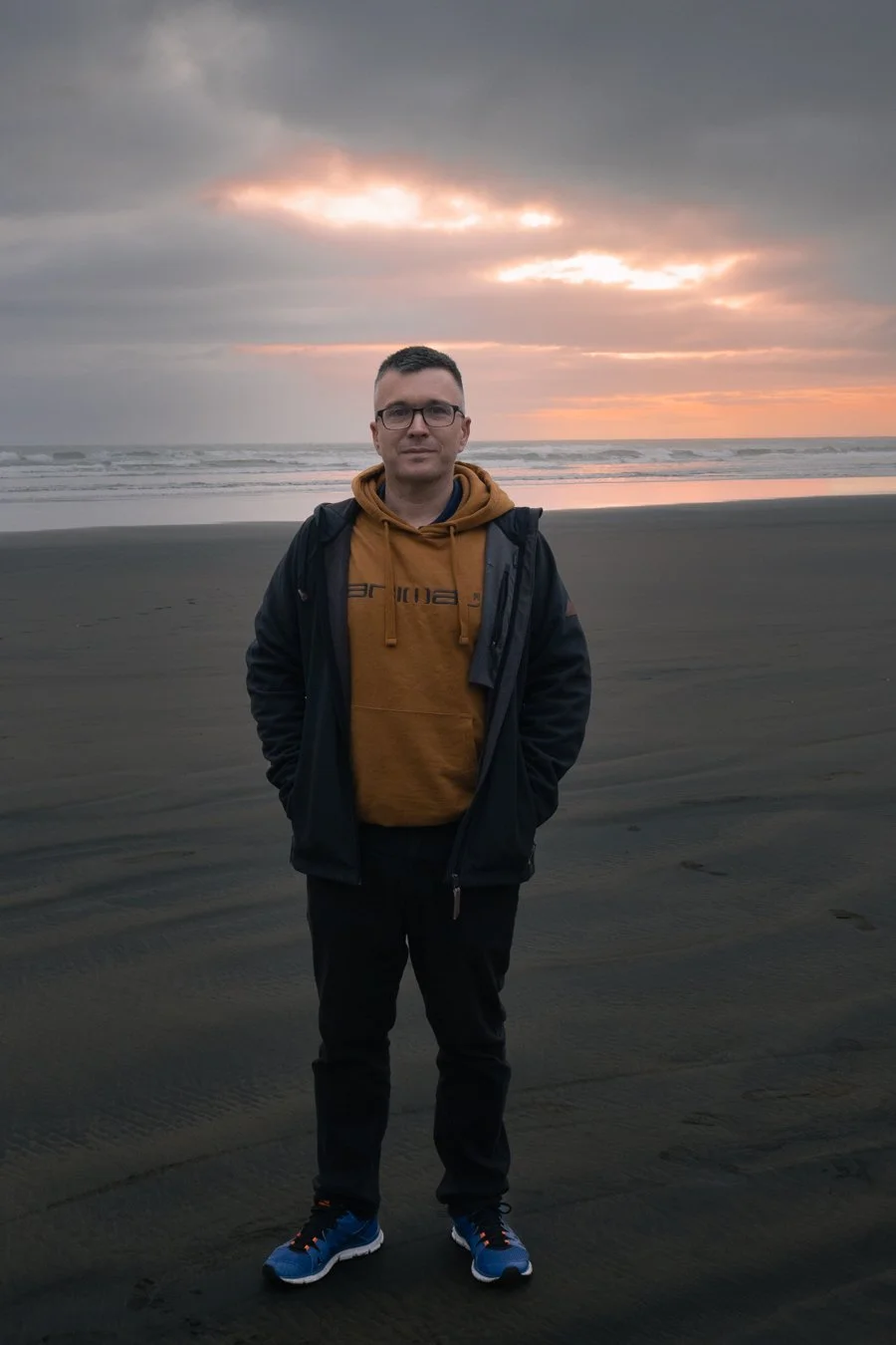 Man standing on a dark sandy beach during sunset, wearing glasses, a mustard hoodie, a black jacket, black pants, and blue sneakers.