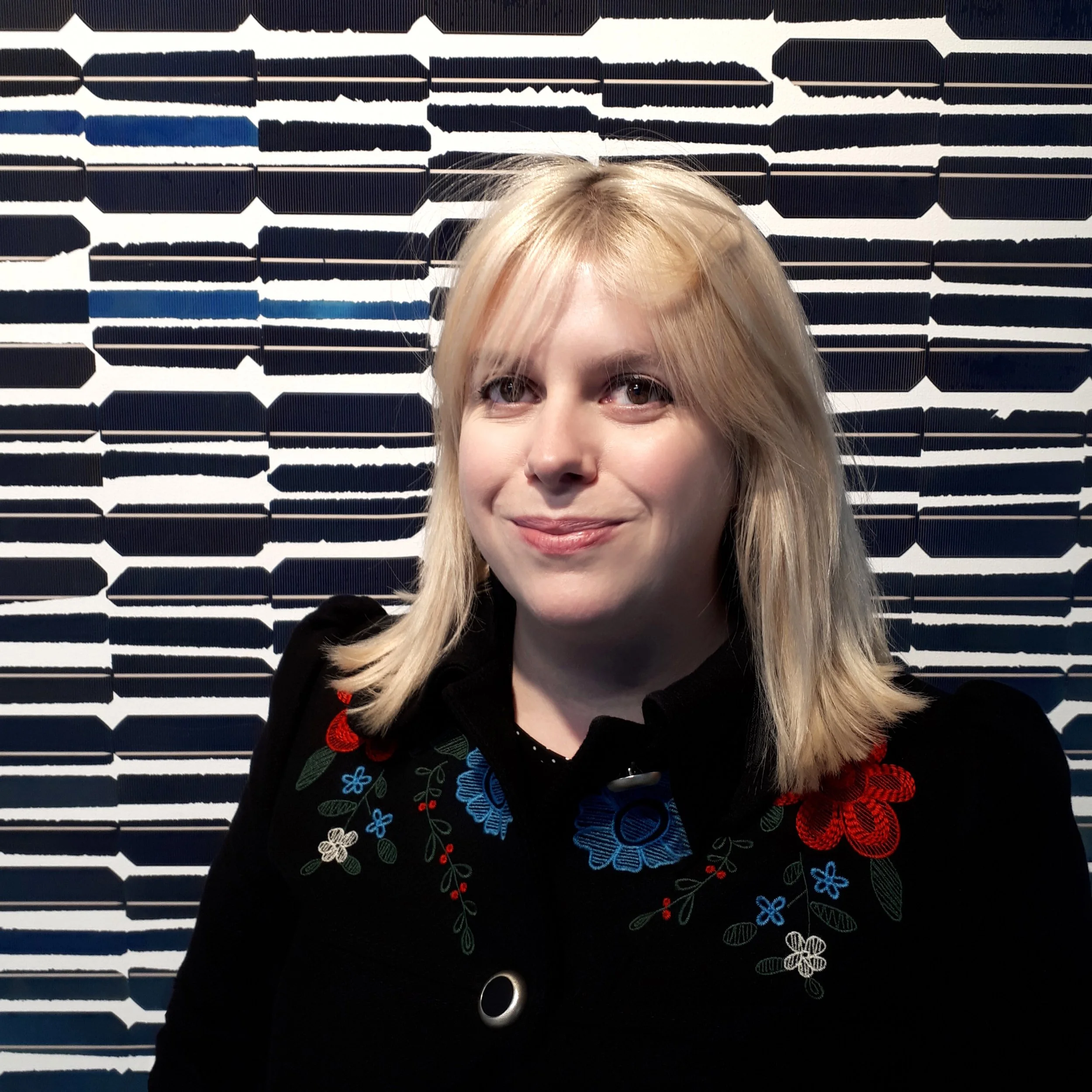 Portrait of a woman with blonde hair standing in front of a patterned black and white artwork.