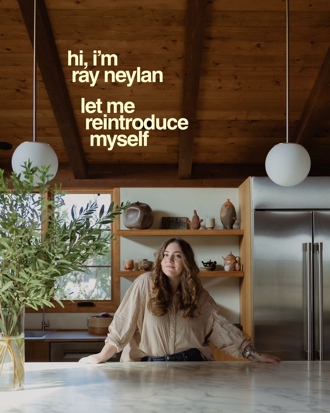 A woman with light skin and wavy brown hair sitting at a kitchen island, with a wooden ceiling and shelves behind her, and text that reads, 'hi, i'm ray neyln let me reintroduce myself.'