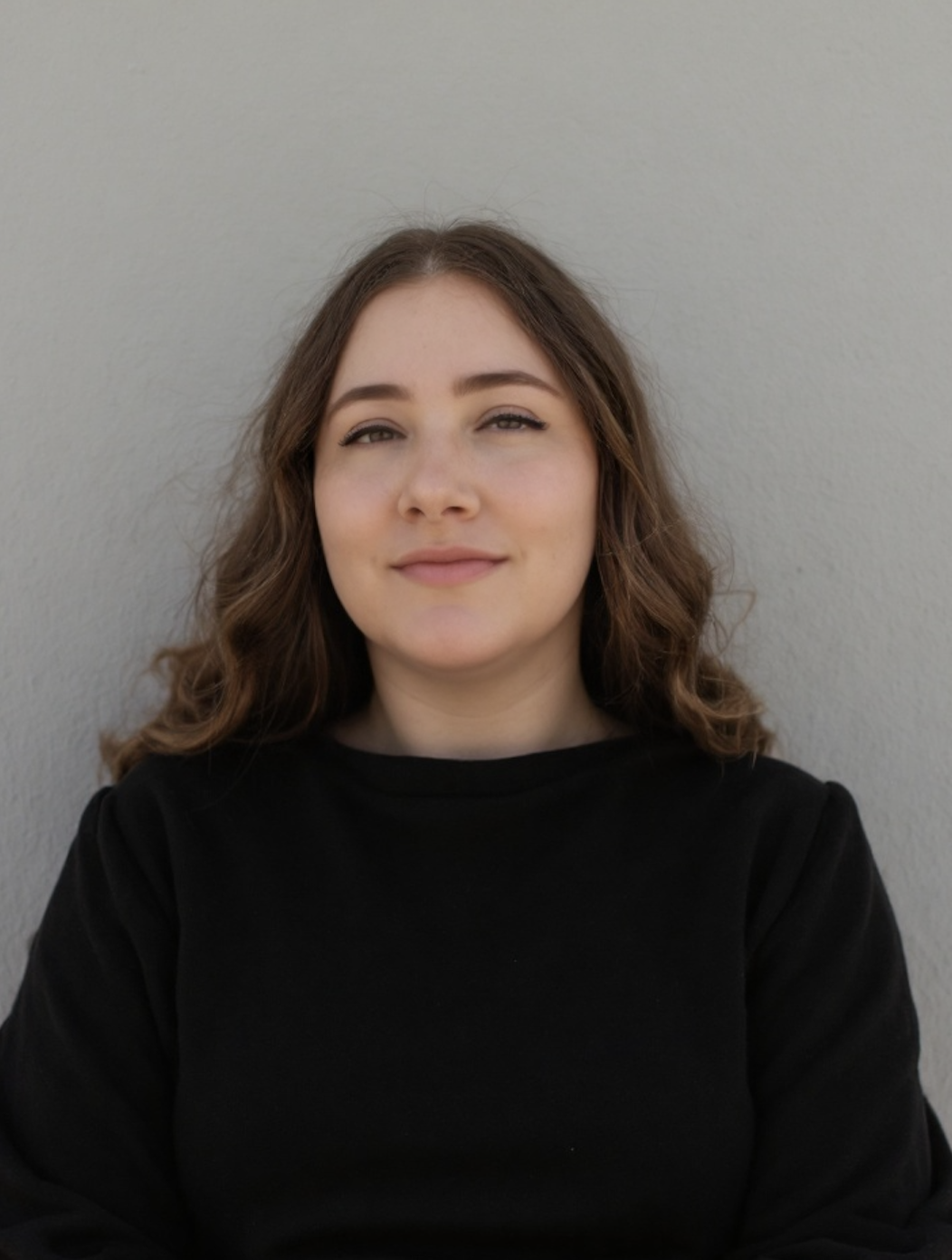 A young woman with shoulder-length curly brown hair, wearing a black shirt, smiling subtly while standing against a plain gray background.
