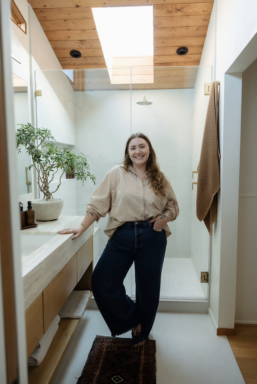 A woman standing in a bathroom, smiling, with a glass shower enclosure behind her, a potted plant on the vanity, and a brown towel hanging on a hook on the wall.