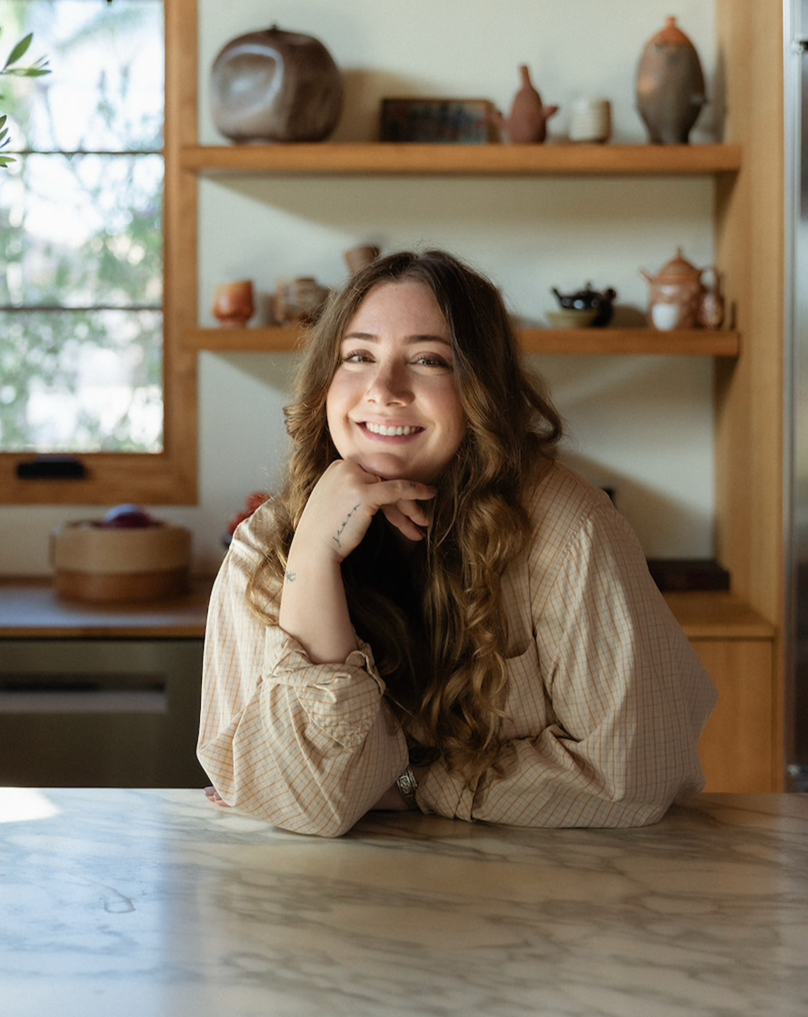 A smiling woman with long, wavy brown hair, resting her chin on her hand, sitting at a wooden table in a cozy room with shelves and a window in the background.