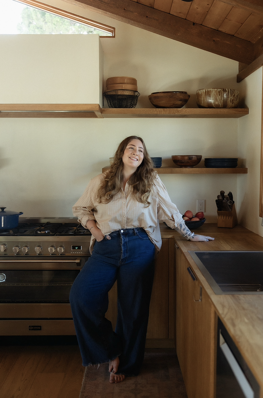 A woman with long curly hair standing in a rustic kitchen, smiling with her hand on the cabinet, wearing a beige checked shirt and dark jeans.