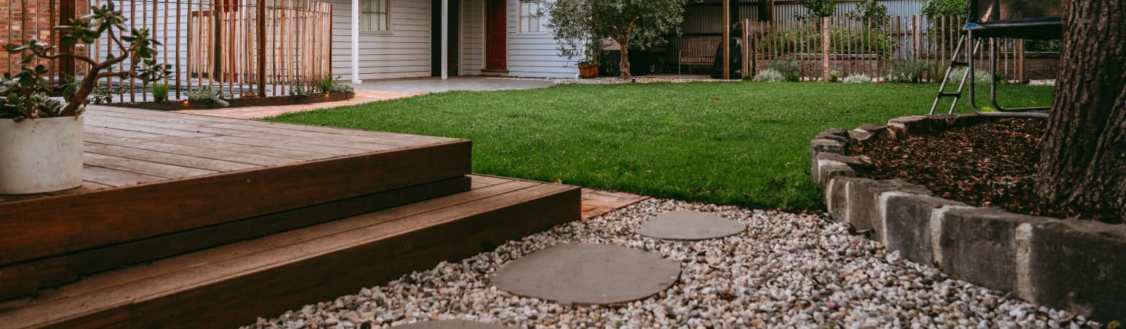 A backyard with a wooden deck, potted plant, green grass lawn, tree, stone pathway, gravel, and a garden bed surrounded by stones.