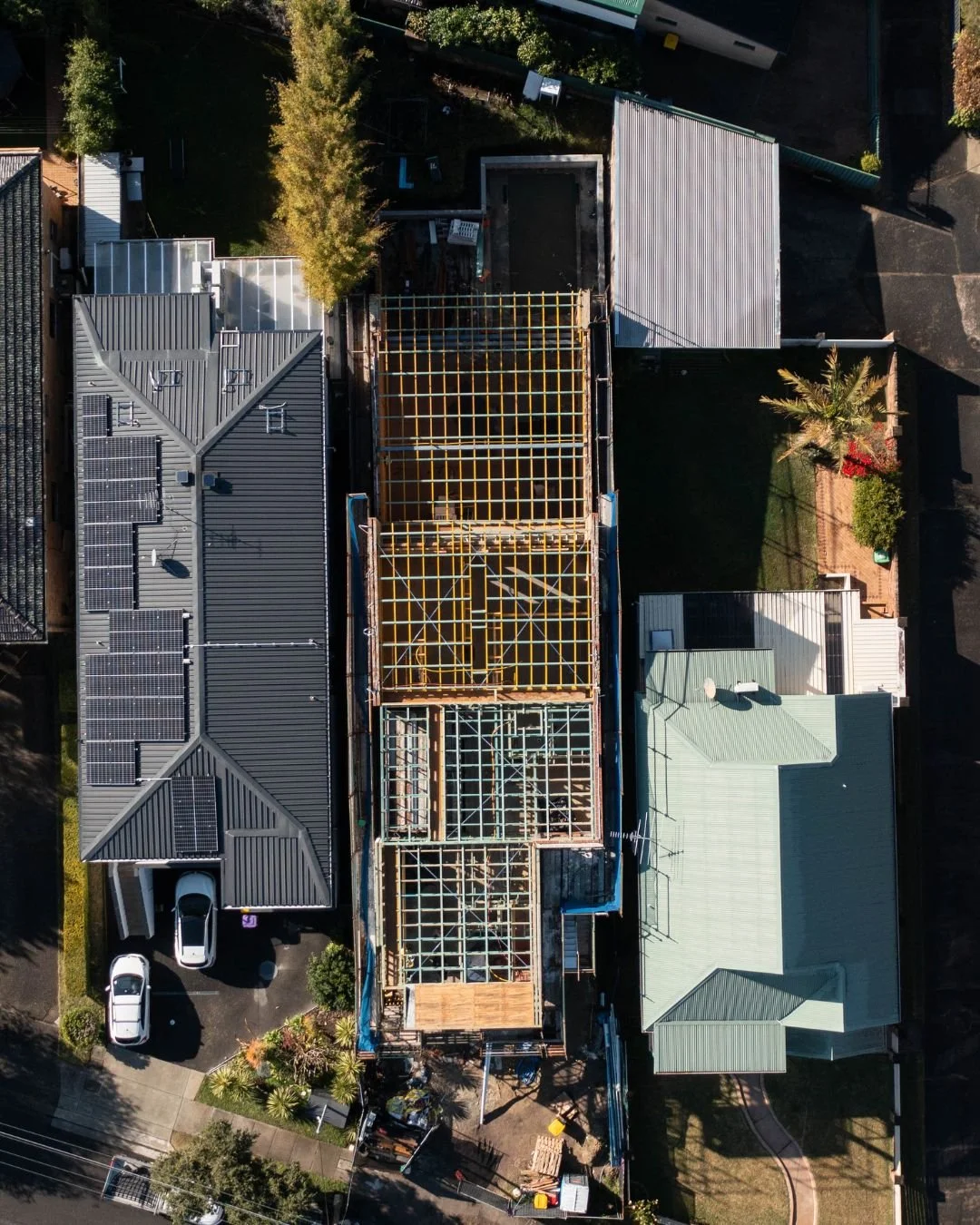 An aerial view of a residential area with a house under construction in the center, surrounded by neighboring houses with solar panels on their roofs, trees, and a driveway with parked cars.