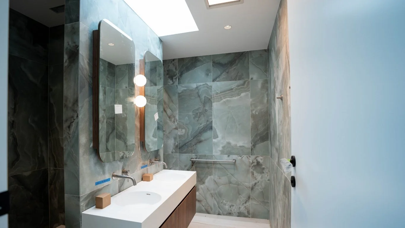 Modern bathroom with gray marble tiles on the walls, a double sink vanity with wooden cabinets, two mirrors, and round light fixtures. There's a towel bar on the side wall and a skylight overhead.