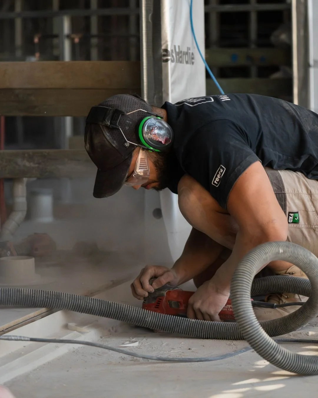 A man working on a construction project, wearing safety goggles, a black cap, headphones around his neck, a black shirt, and tan shorts, using a power saw on the floor amidst dust and construction materials.