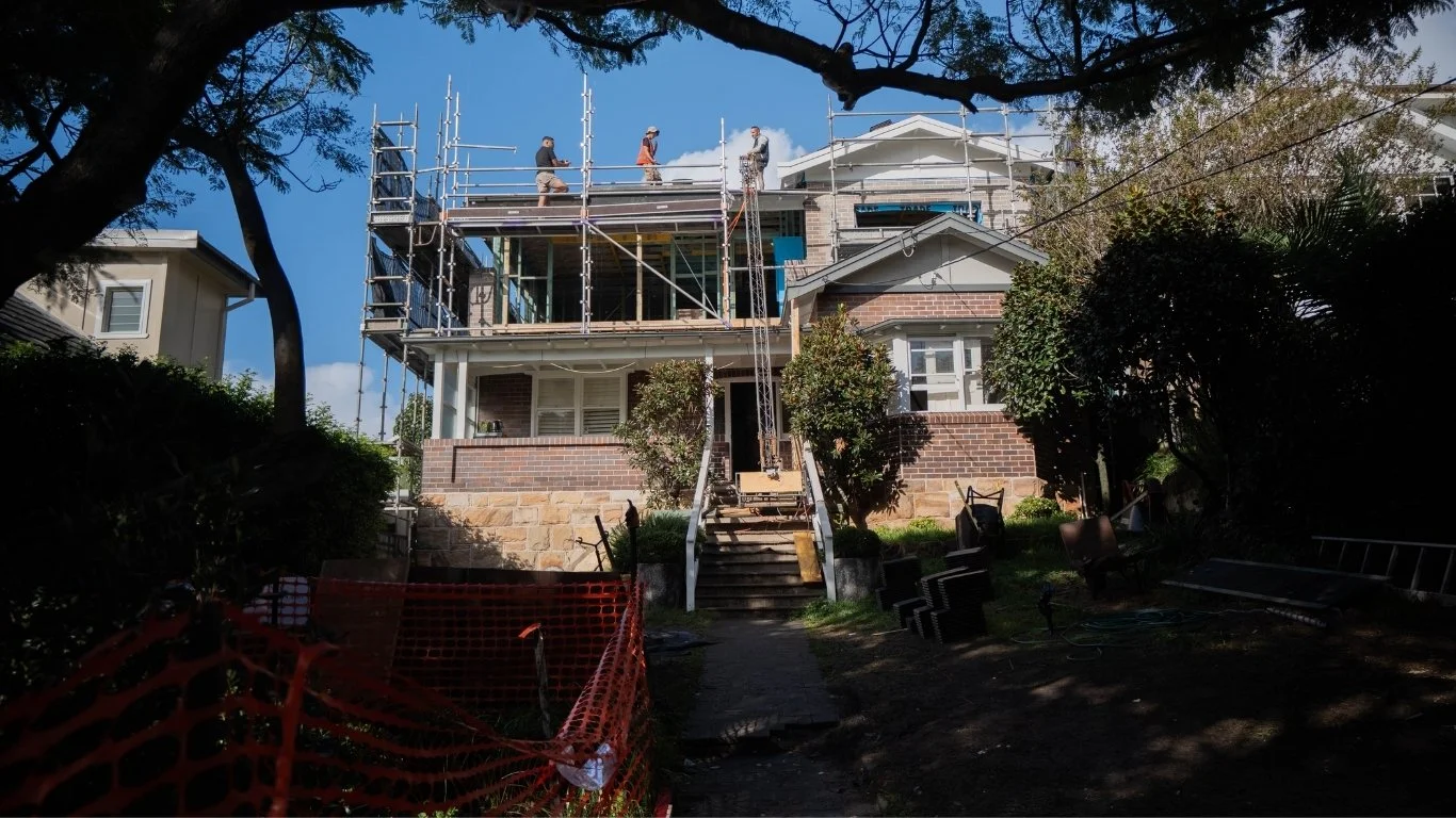 House under construction with scaffolding on the upper level, workers on the scaffolding, and construction materials and tools on the ground.