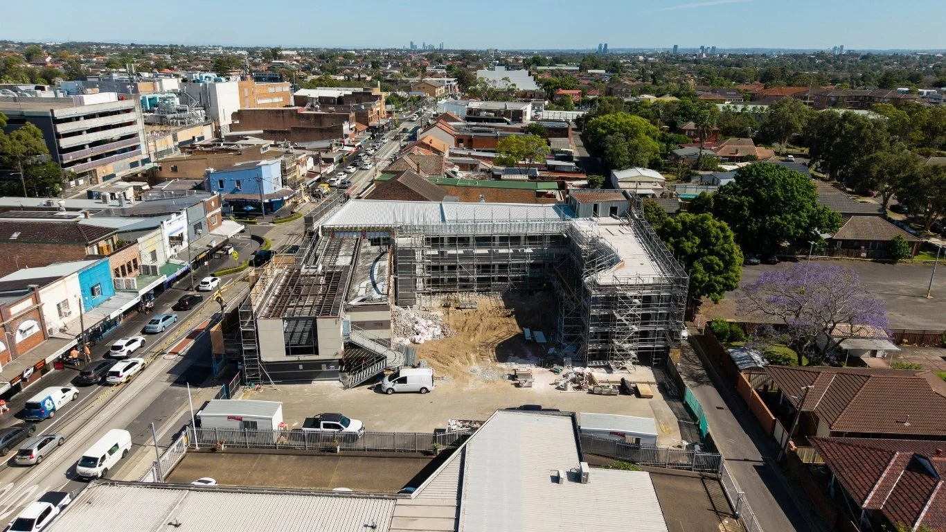 Aerial view of a construction site in an urban area with scaffolding around the building, surrounding streets with parked cars, and residential and commercial buildings in the background.