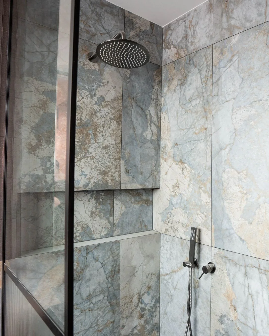 Close-up view of a modern shower area with gray and white marble tiles, a round overhead shower head, and a handheld shower fixture.